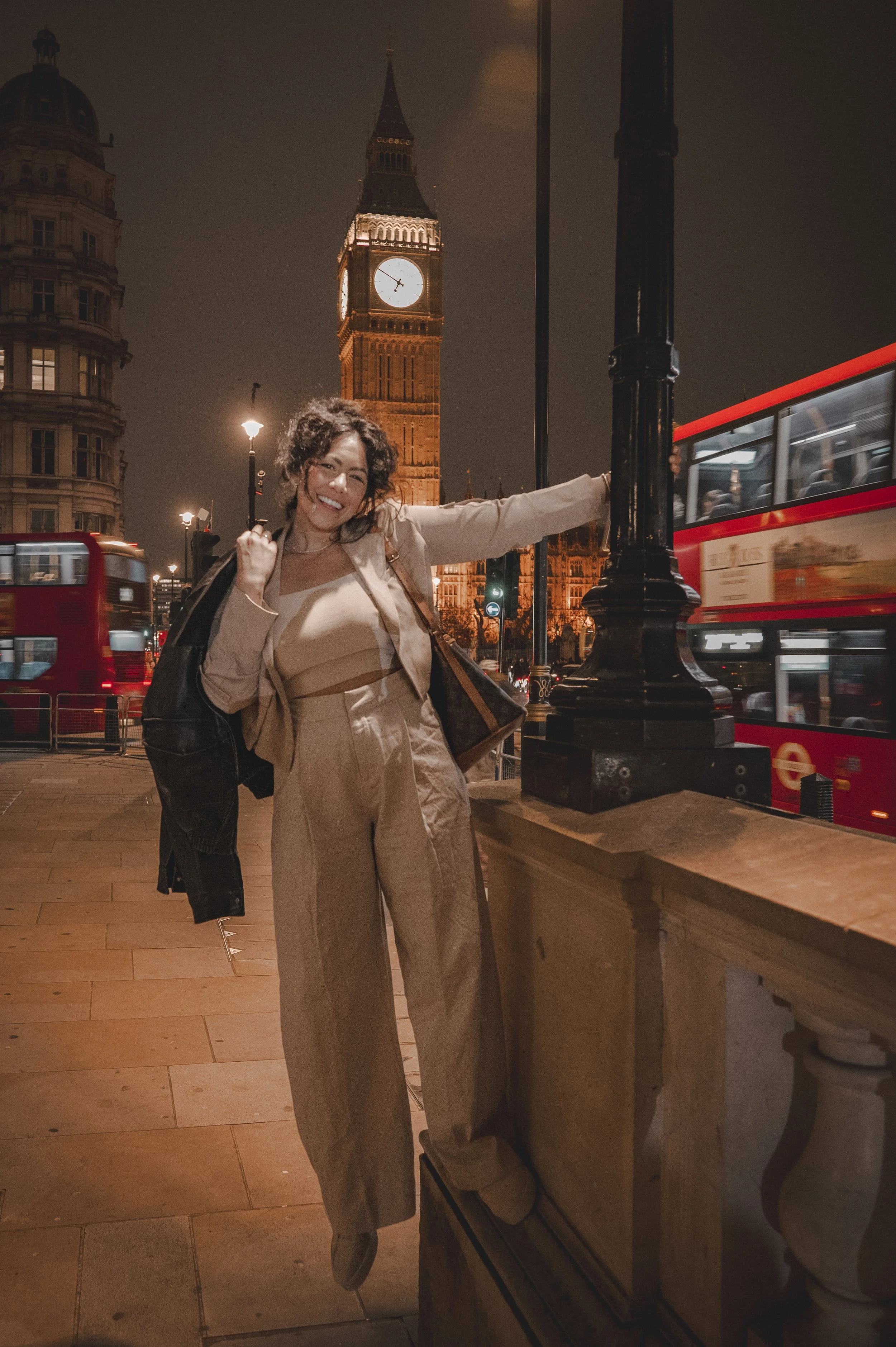 A woman smiling at night in front of Big Ben in London, with red double-decker buses passing by.
