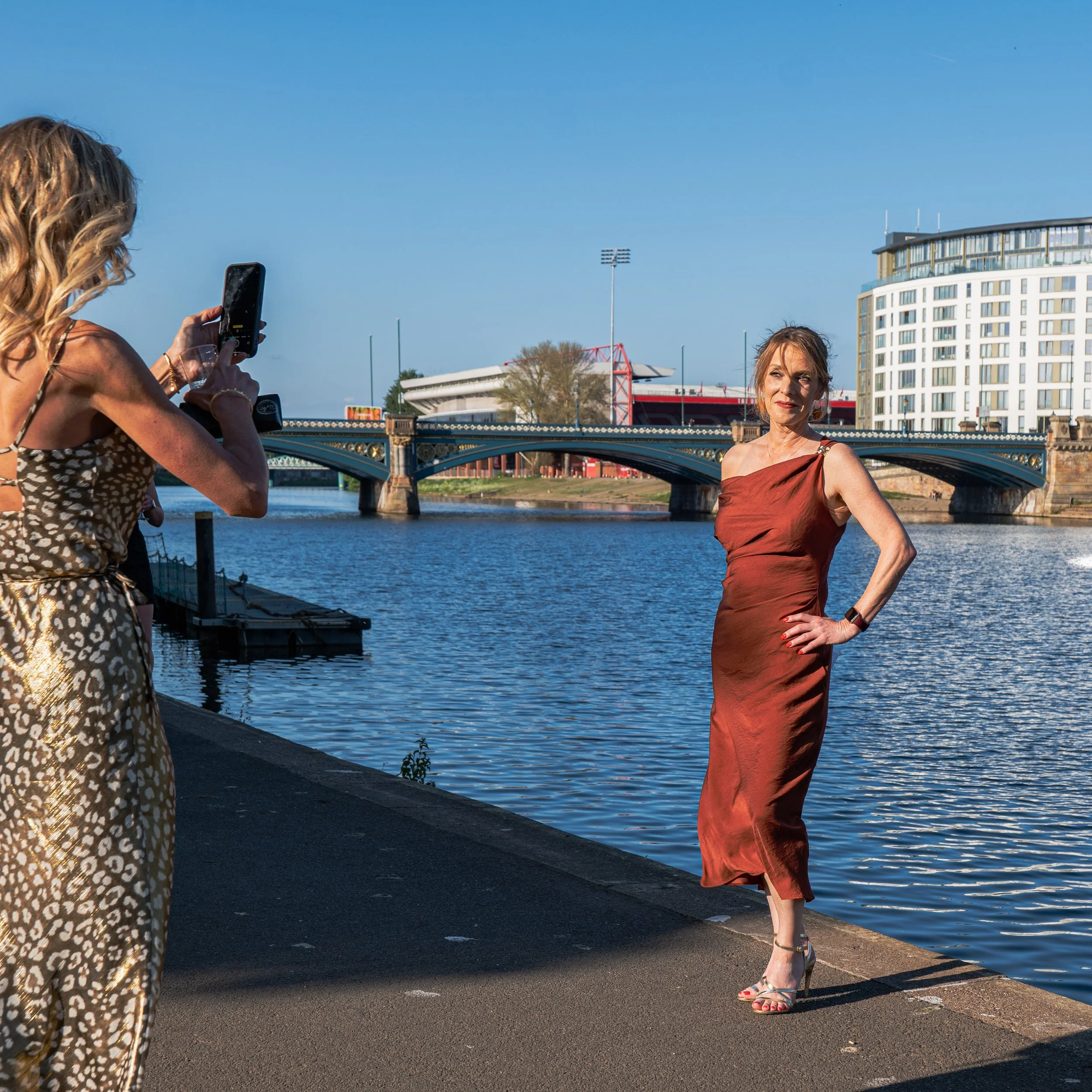 A woman in a red satin dress posing by the water while another woman takes her photo along a riverside promenade.