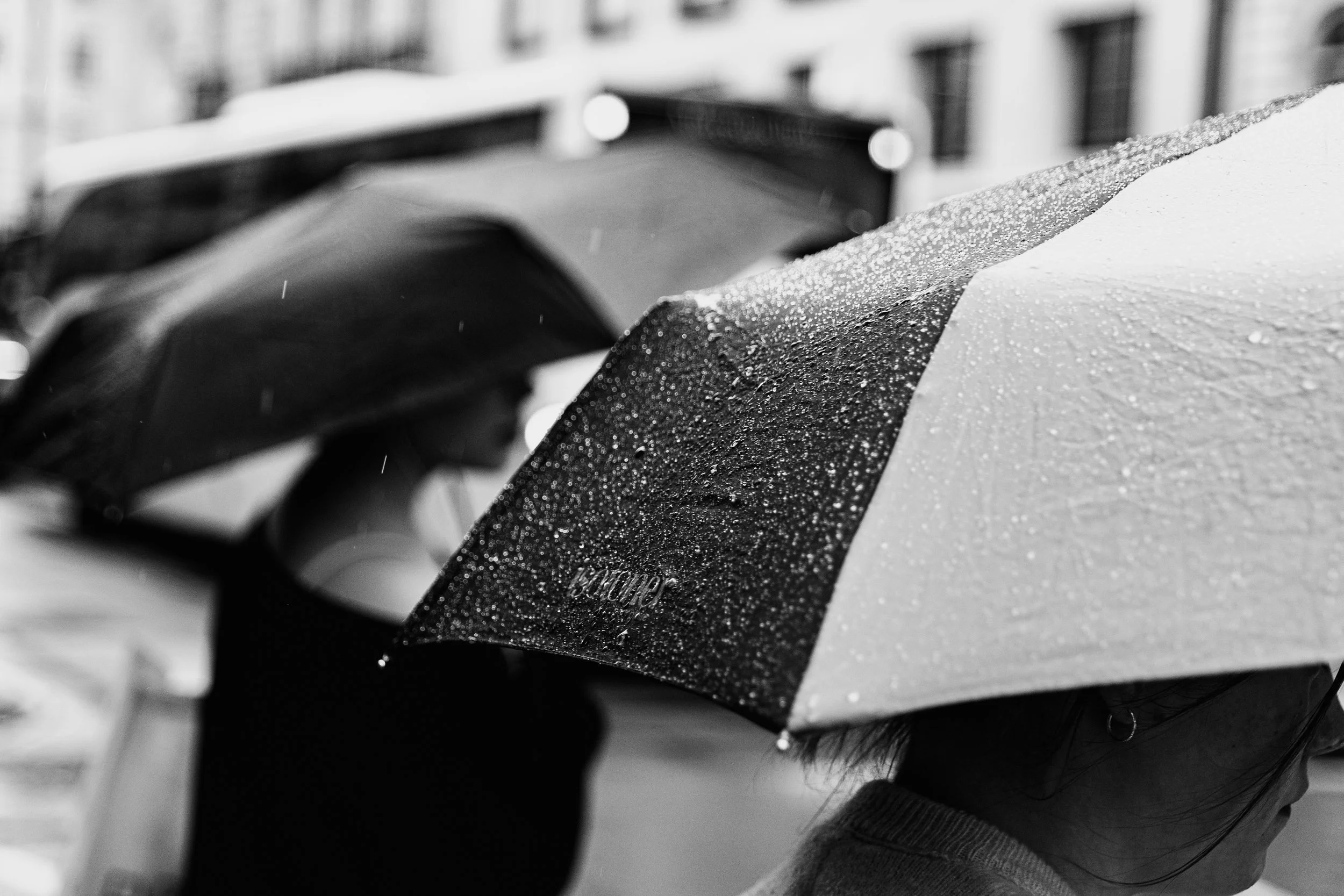 Two people walking with umbrellas in the rain on a city street, one person with a black umbrella and the other with a patterned umbrella, with wet surfaces and blurred buildings in the background.