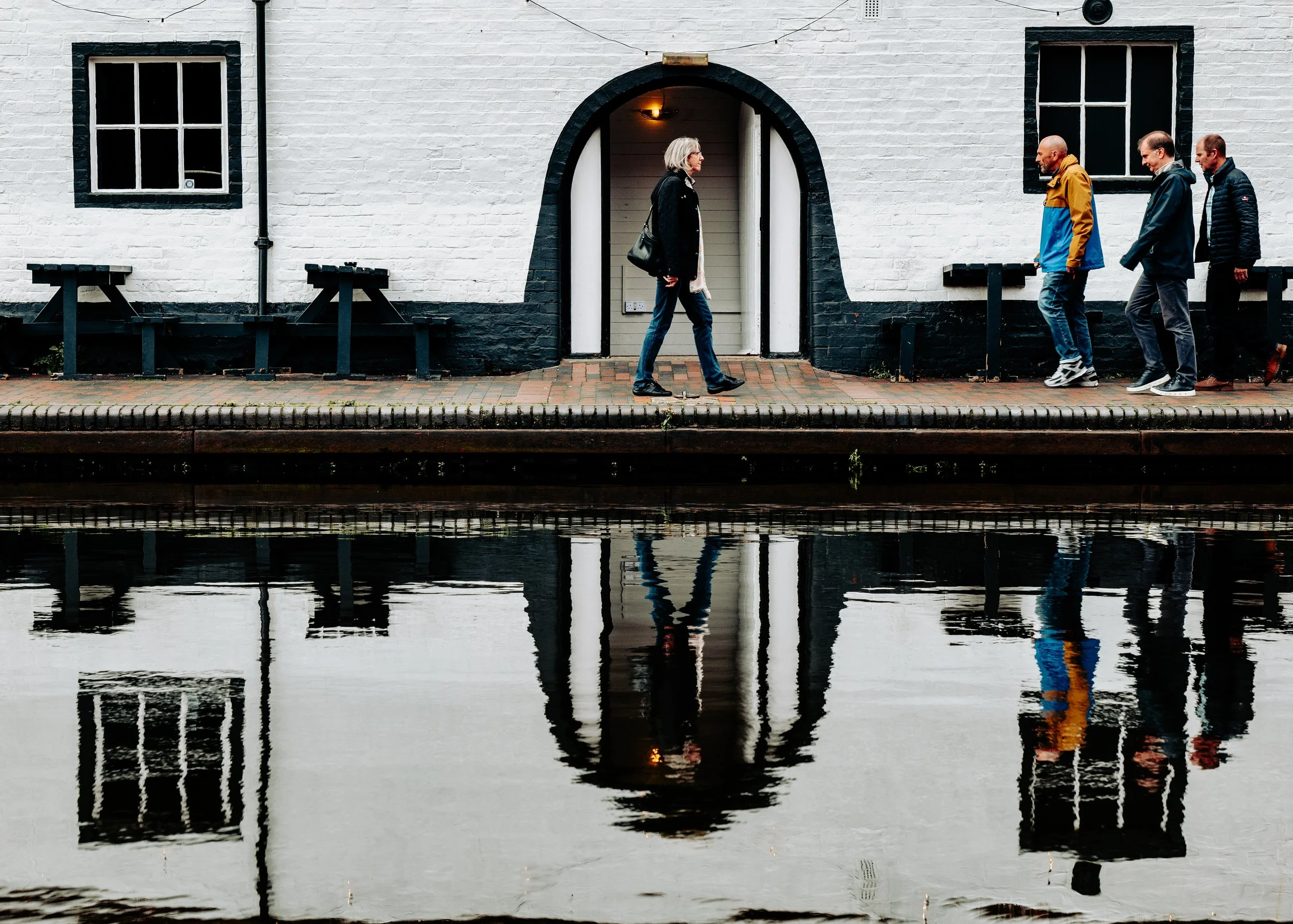 A woman walking along a sidewalk near a canal, with her reflection visible in the water. Four men are walking behind her, also reflected in the canal. The background features a white building with black window frames and benches.