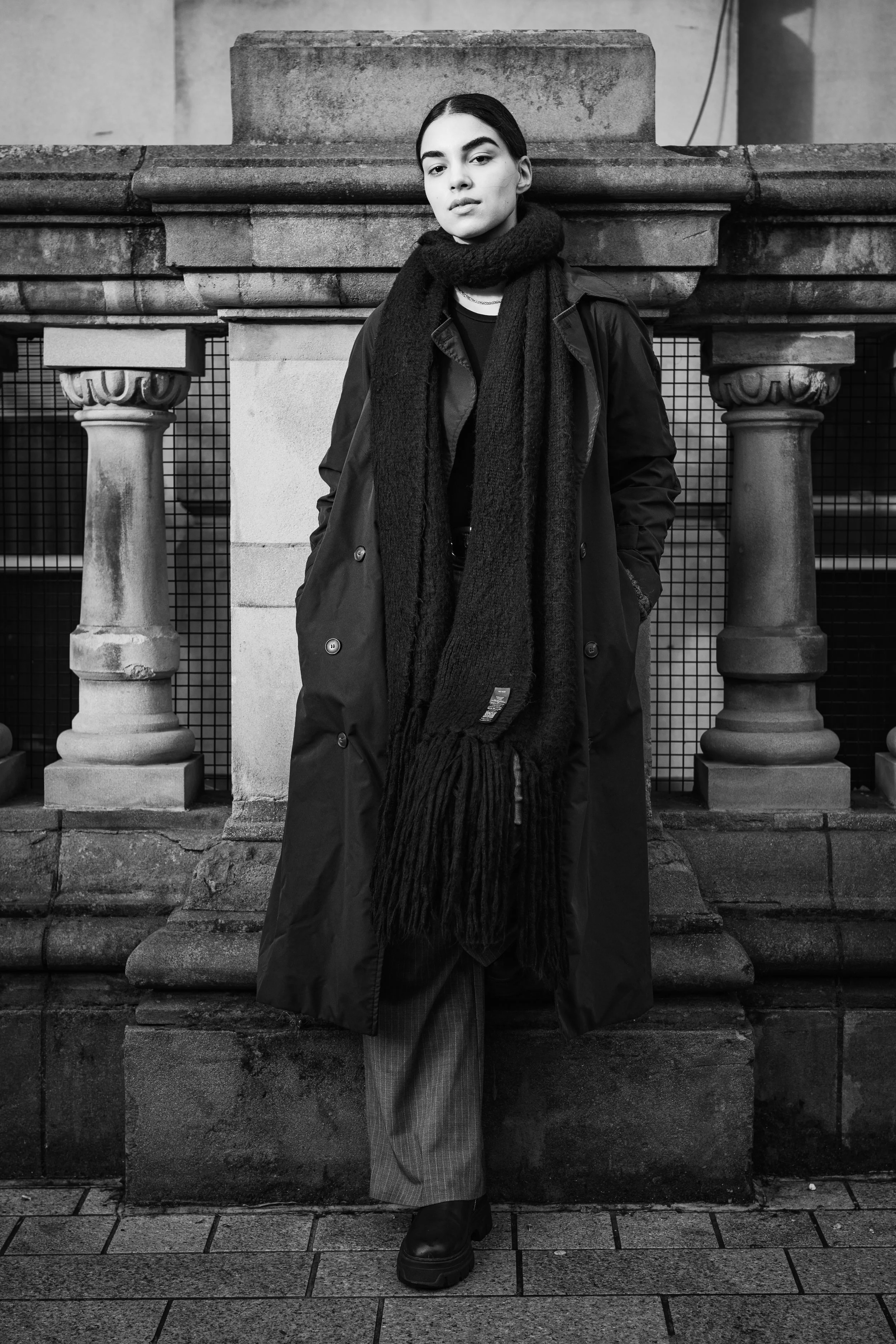 A woman with dark hair in a low ponytail wearing a long coat, a large scarf, and pants standing in front of an old stone structure with columns on an urban sidewalk in black and white.