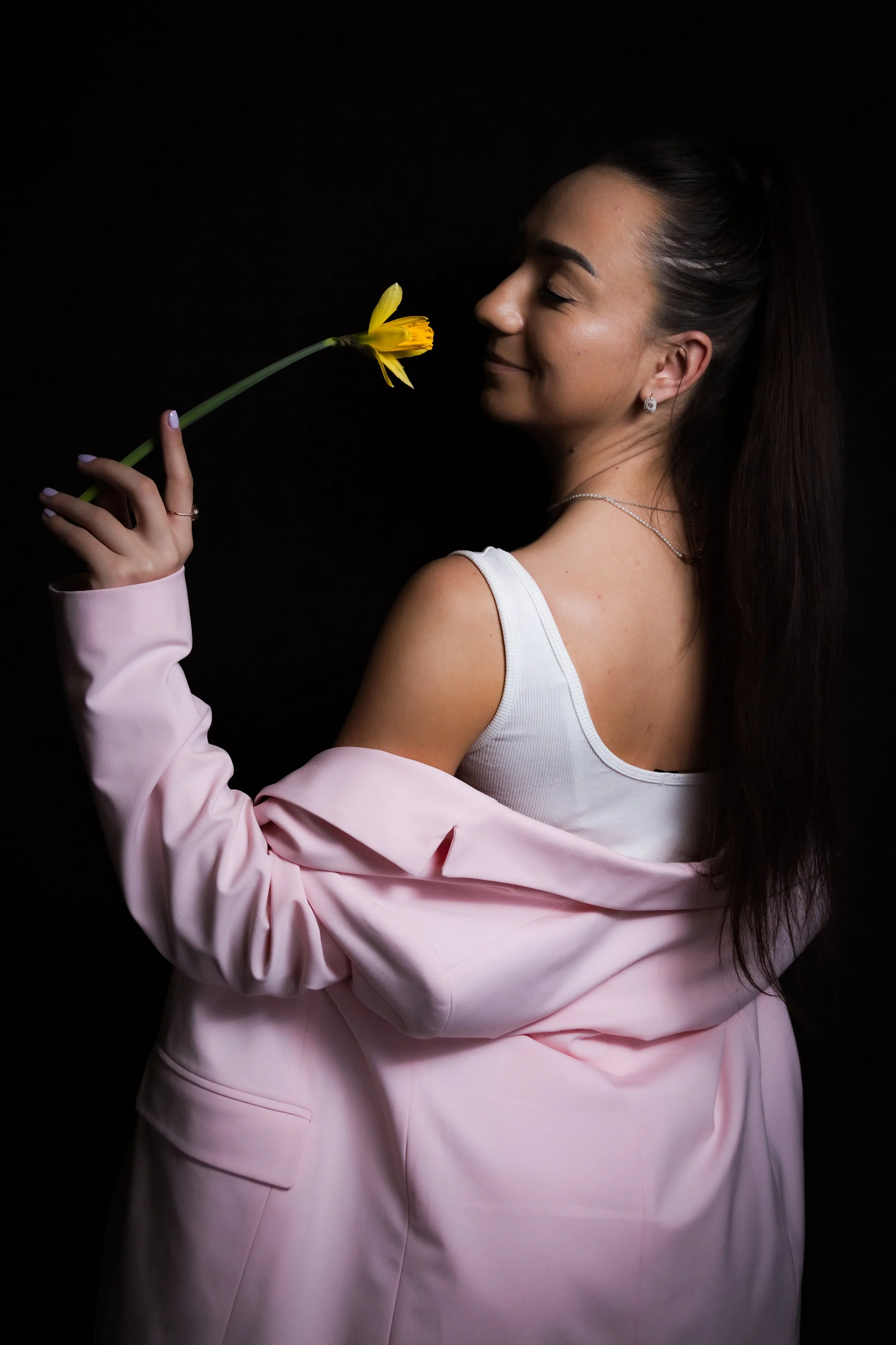 Young woman with long dark hair, wearing a white tank top and pink blazer, holding a yellow flower close to her face with a gentle smile, against a black background.