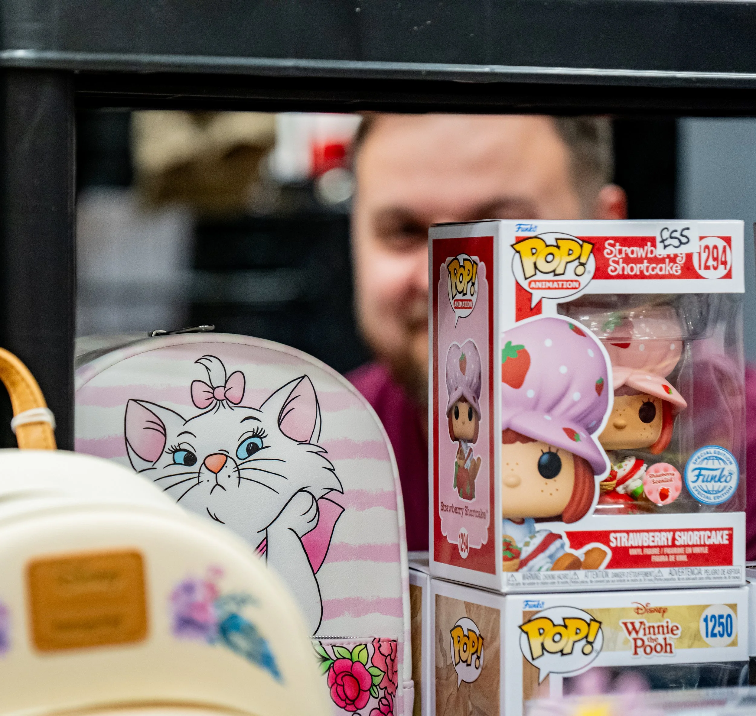 A man is partially visible behind a display shelf with cartoon character merchandise, including a boxed Funko Pop! figure of Strawberry Shortcake and a figurine of Winnie the Pooh. There is also a white bag with a drawing of a smiling, pink bow-tied 
