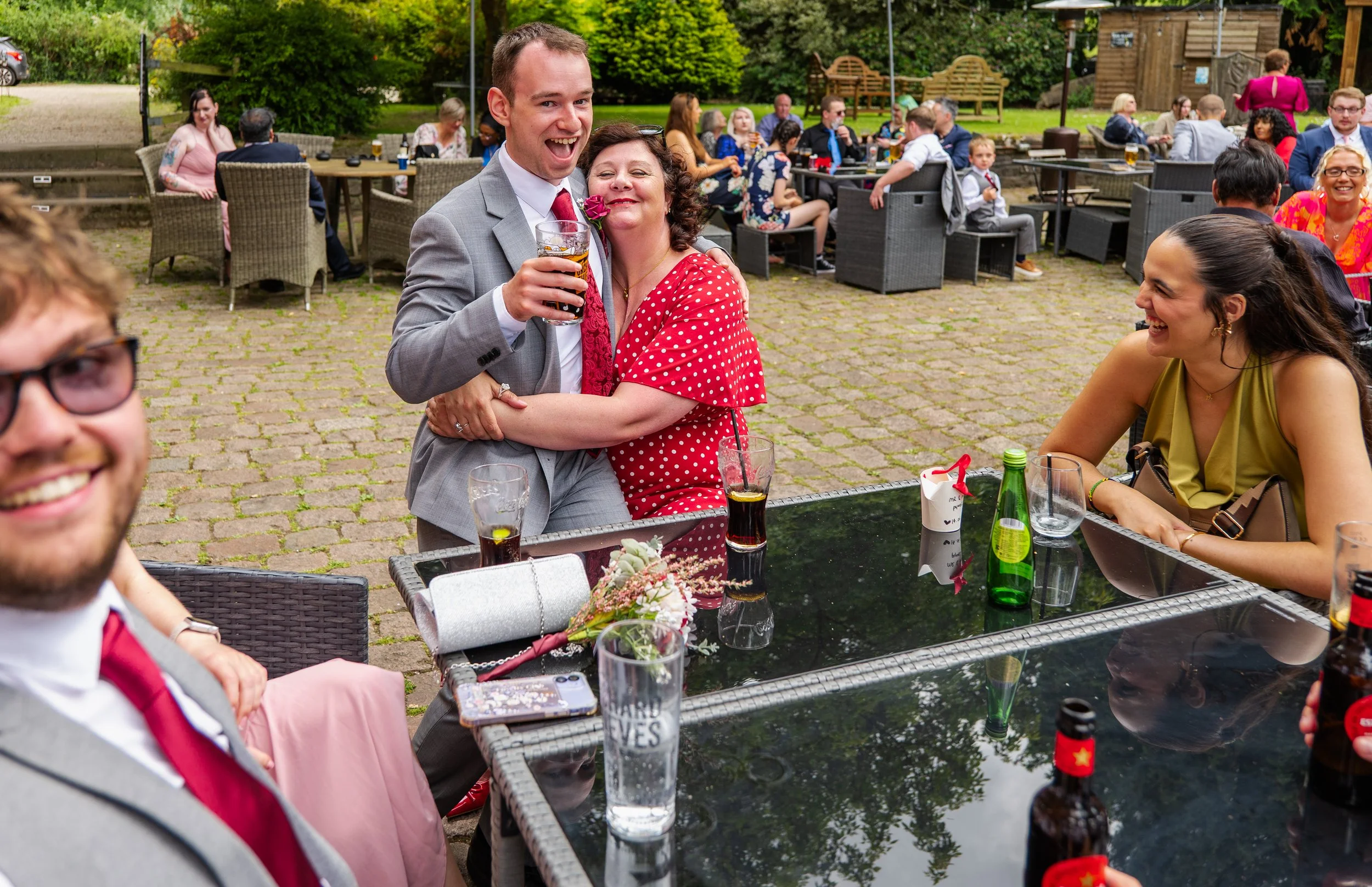 Group of people enjoying outdoor celebration, with a woman hugging a man in a suit, smiles, and glasses, at a patio with drinks and flowers.