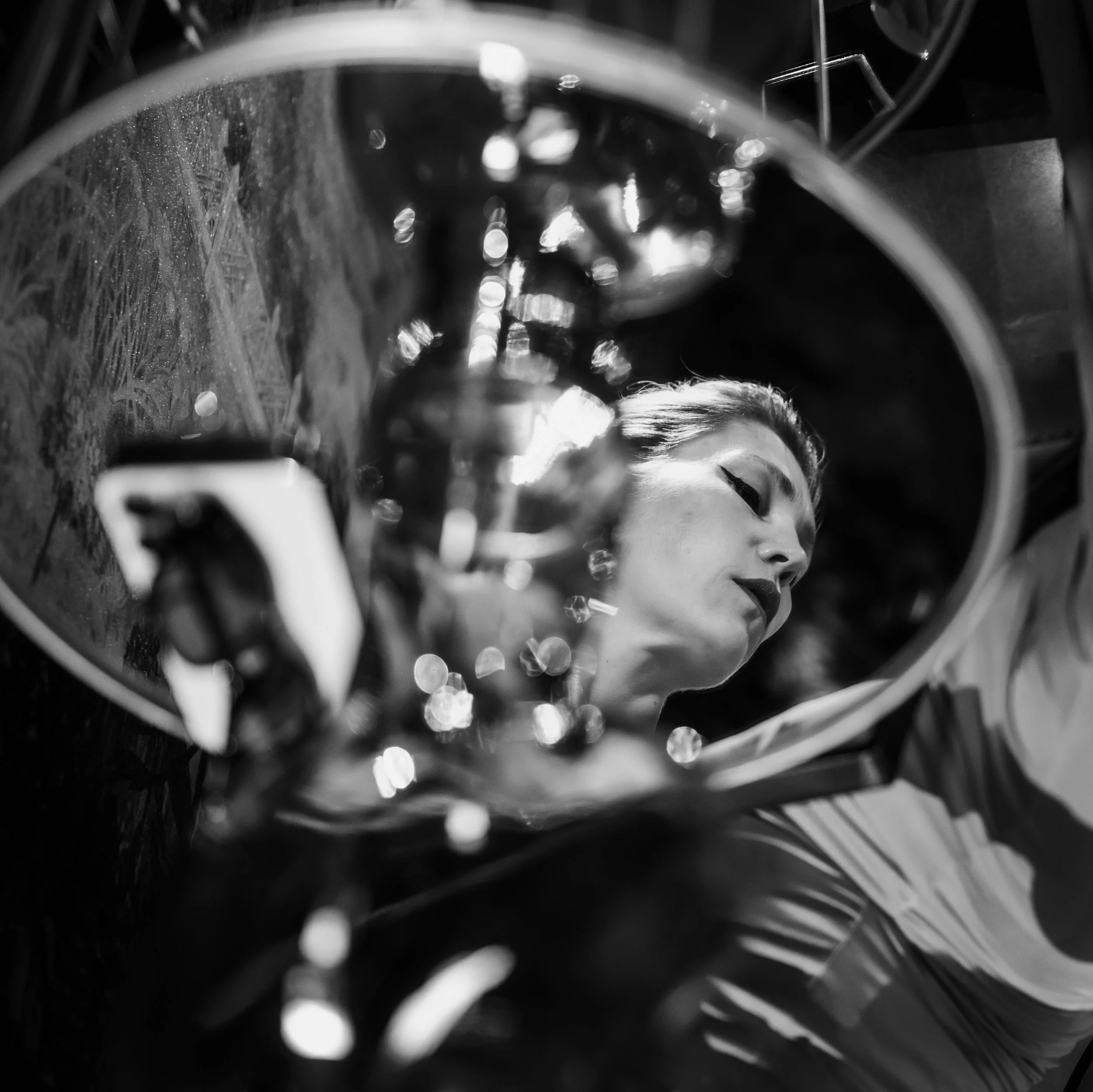 A black and white photo of a woman with makeup and styled hair, seen through a reflective surface or mirror, with blurred lights in the foreground.