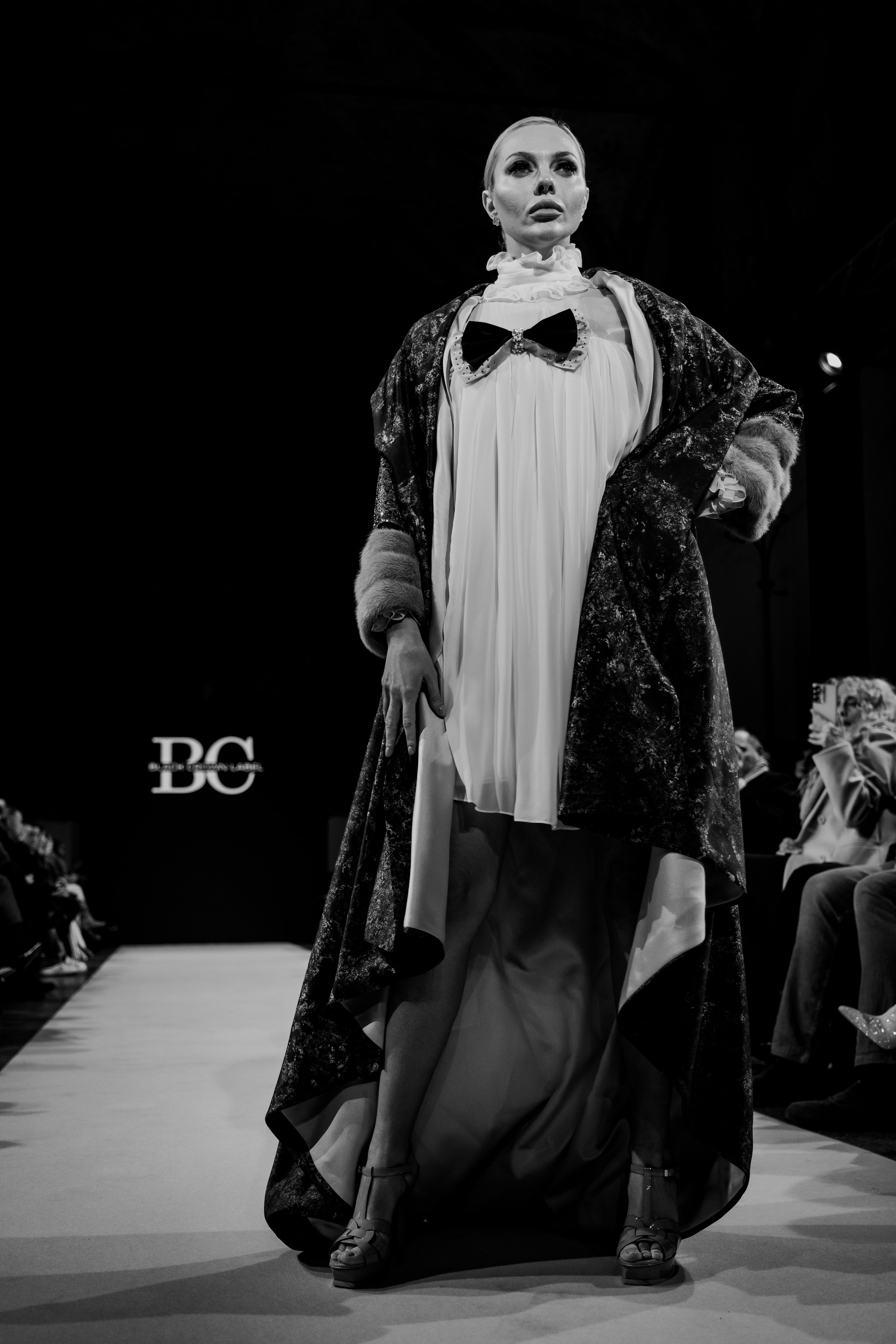 Black and white photo of a fashion model walking on a runway, wearing an elegant long coat over a pleated dress with a large bow, high heels, and makeup, with audience members on the side watching.