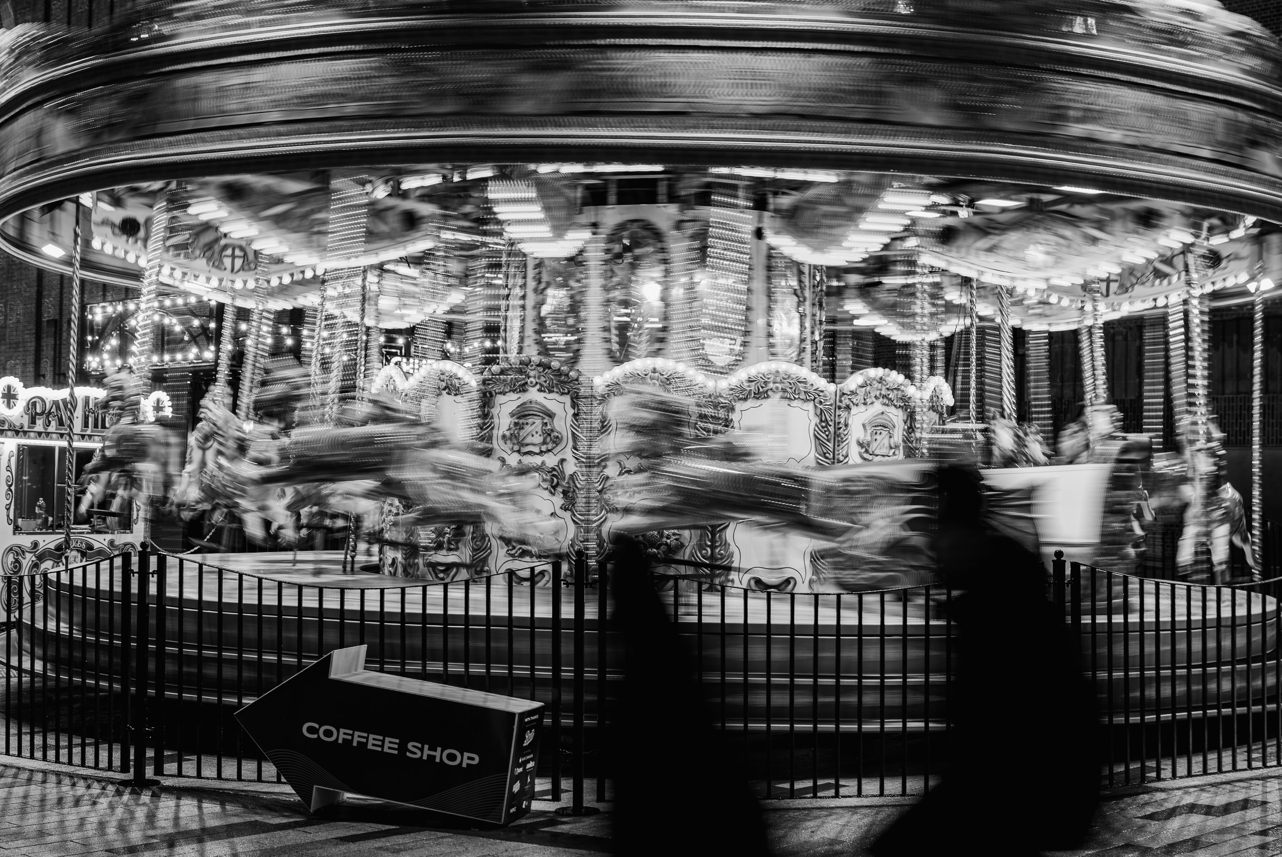 Black and white photo of a carousel at night with blurred motion from spinning. People are walking past the carousel, and a sign pointing to a coffee shop is in front.