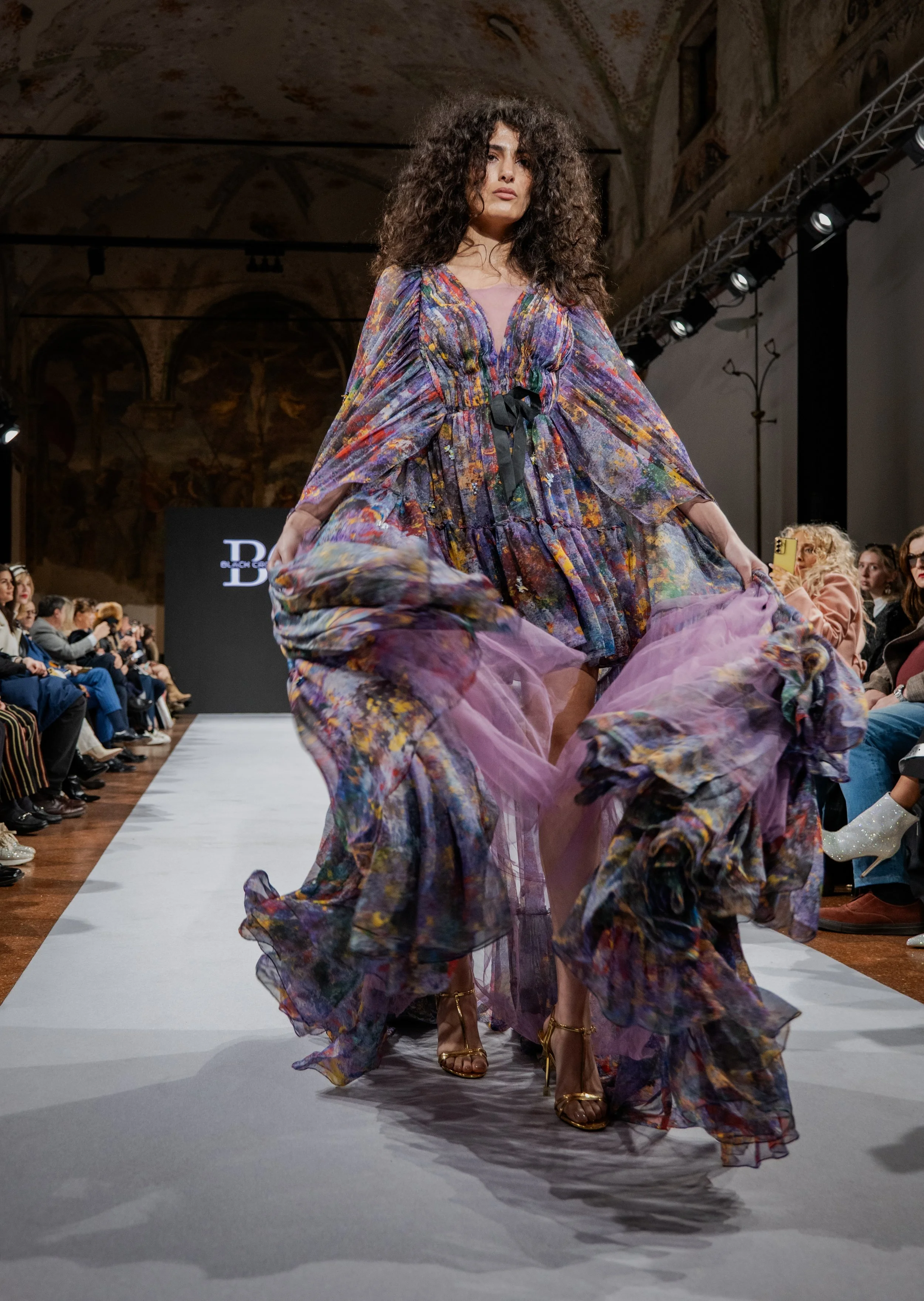 Model walking down a runway wearing a colorful, flowing, floral-patterned dress with purple tulle accents at a fashion show.