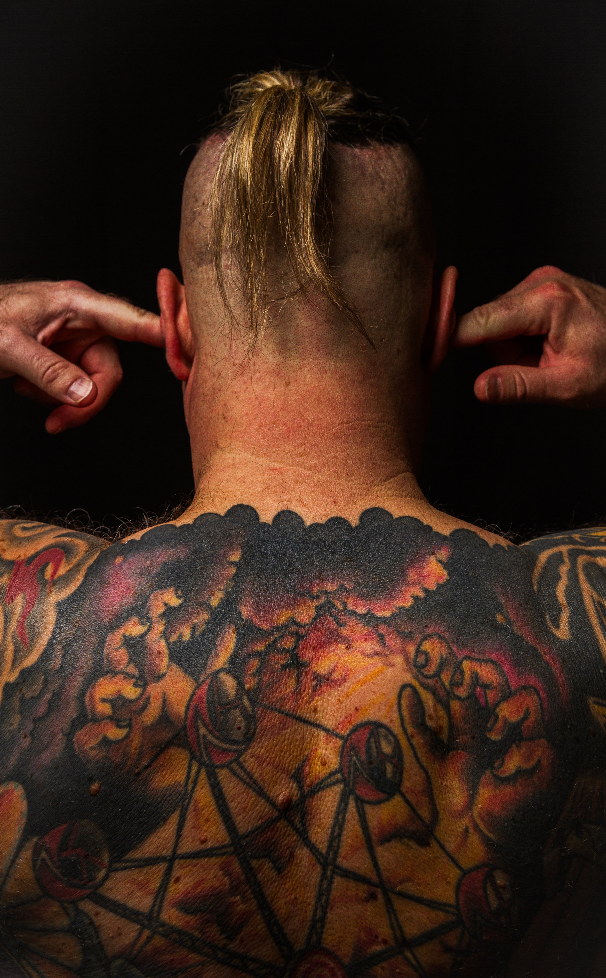 Person with tattoos on their back and neck, pressing fingers into ears, viewed from behind.