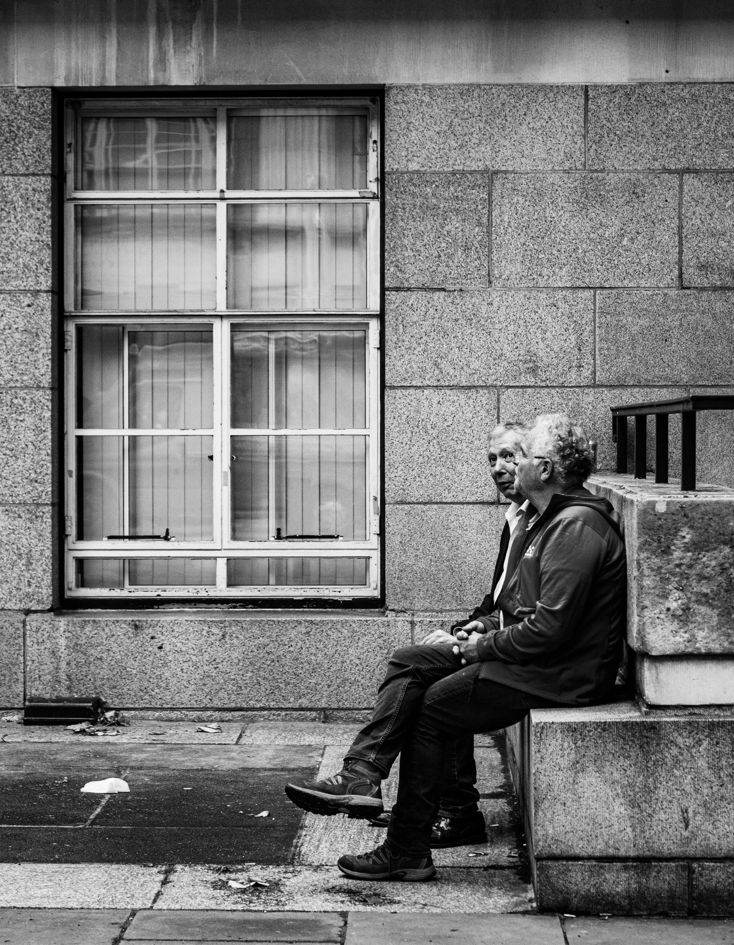 Two men sit on a stone bench on the sidewalk in front of a building with a large window, engaging in conversation.