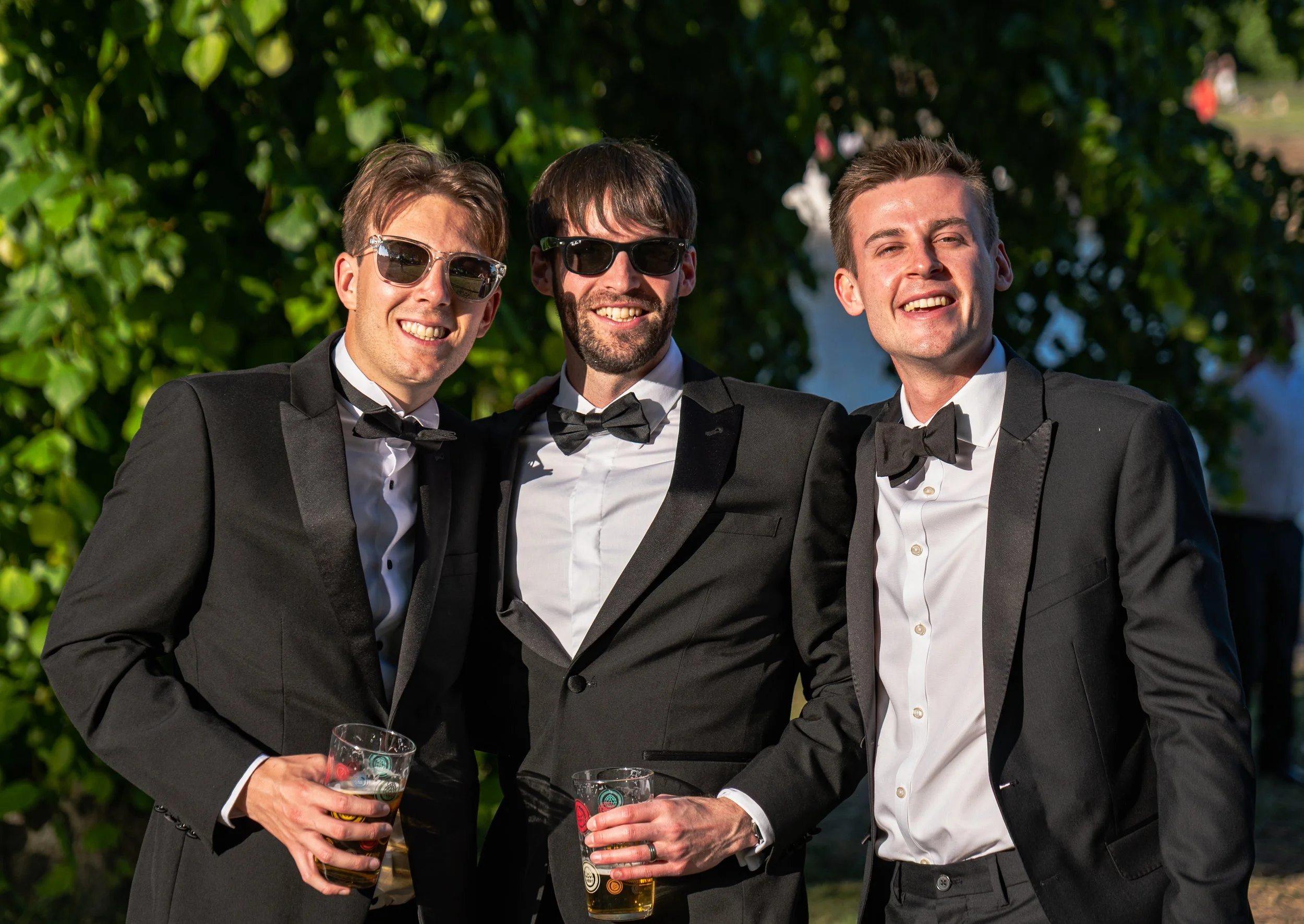 Three young men in tuxedos and sunglasses smiling while holding drinks at an outdoor event.