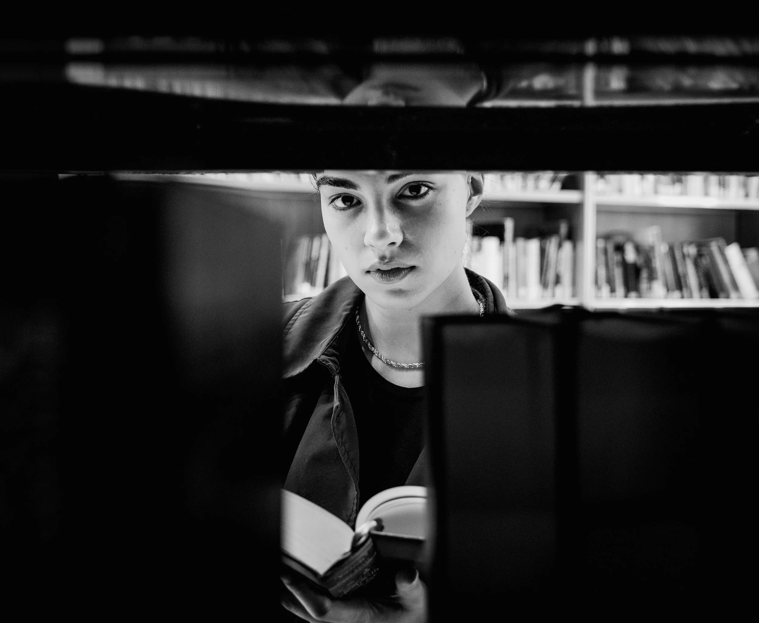 A young woman with short hair and a necklace looking through a gap between bookshelves in a library, black and white photo.