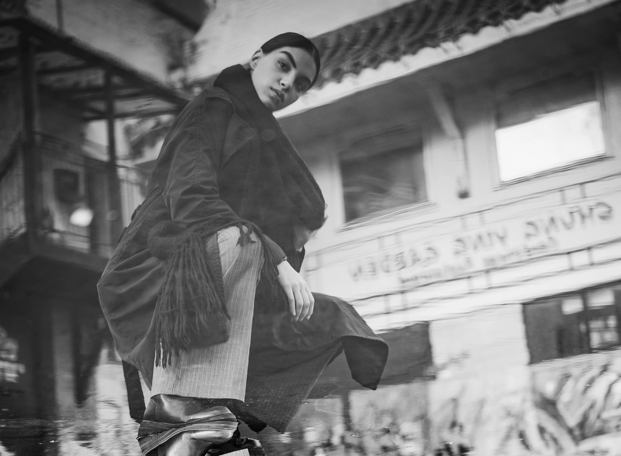 A young woman with dark hair and a serious expression crouches on a street in black and white. She is dressed in a long coat, scarf, and striped pants, with boots. The background has a building with a sign, windows, and some structural elements.