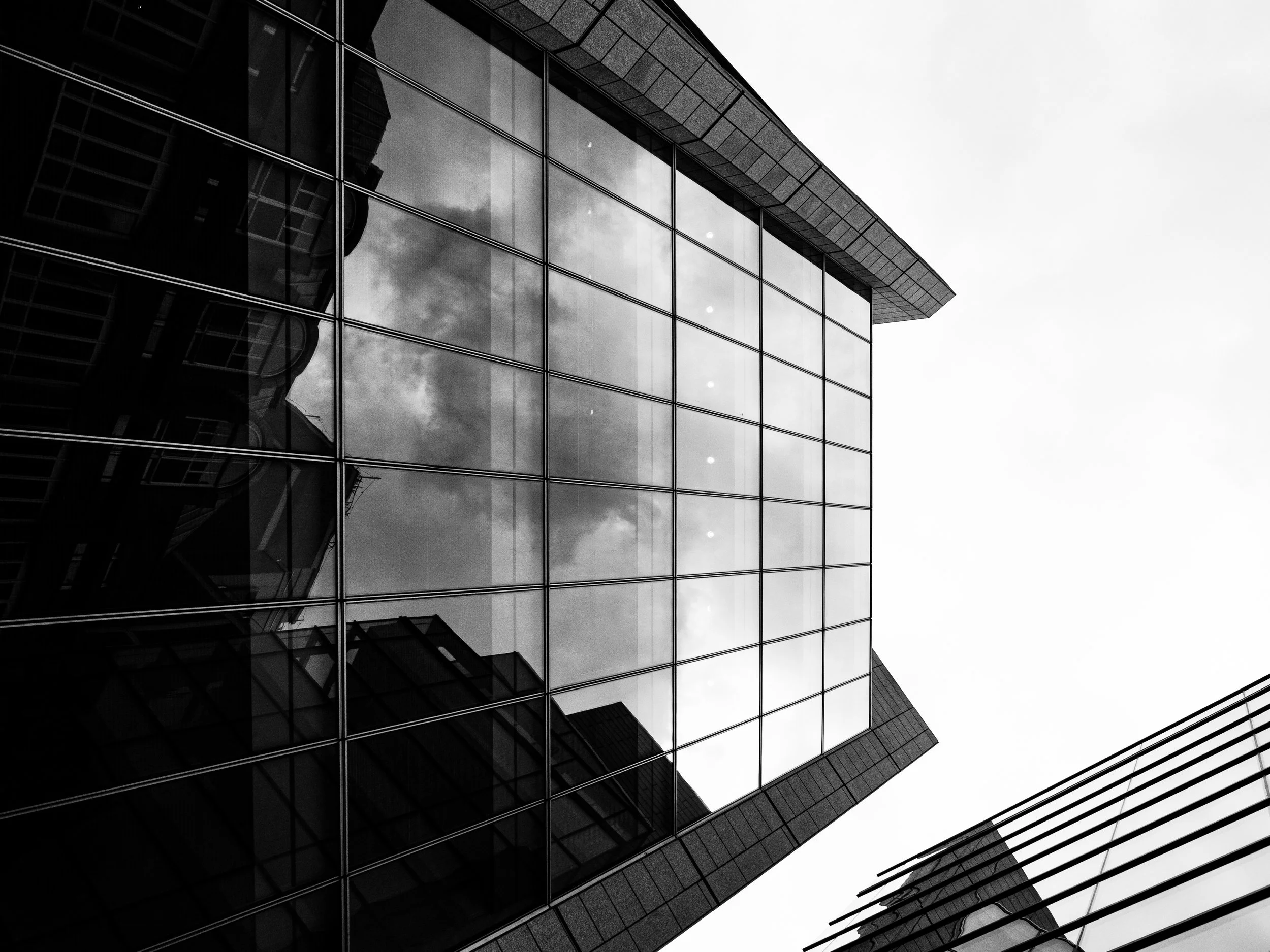 Black and white photo of a modern glass building with reflections of nearby structures and cloudy sky.