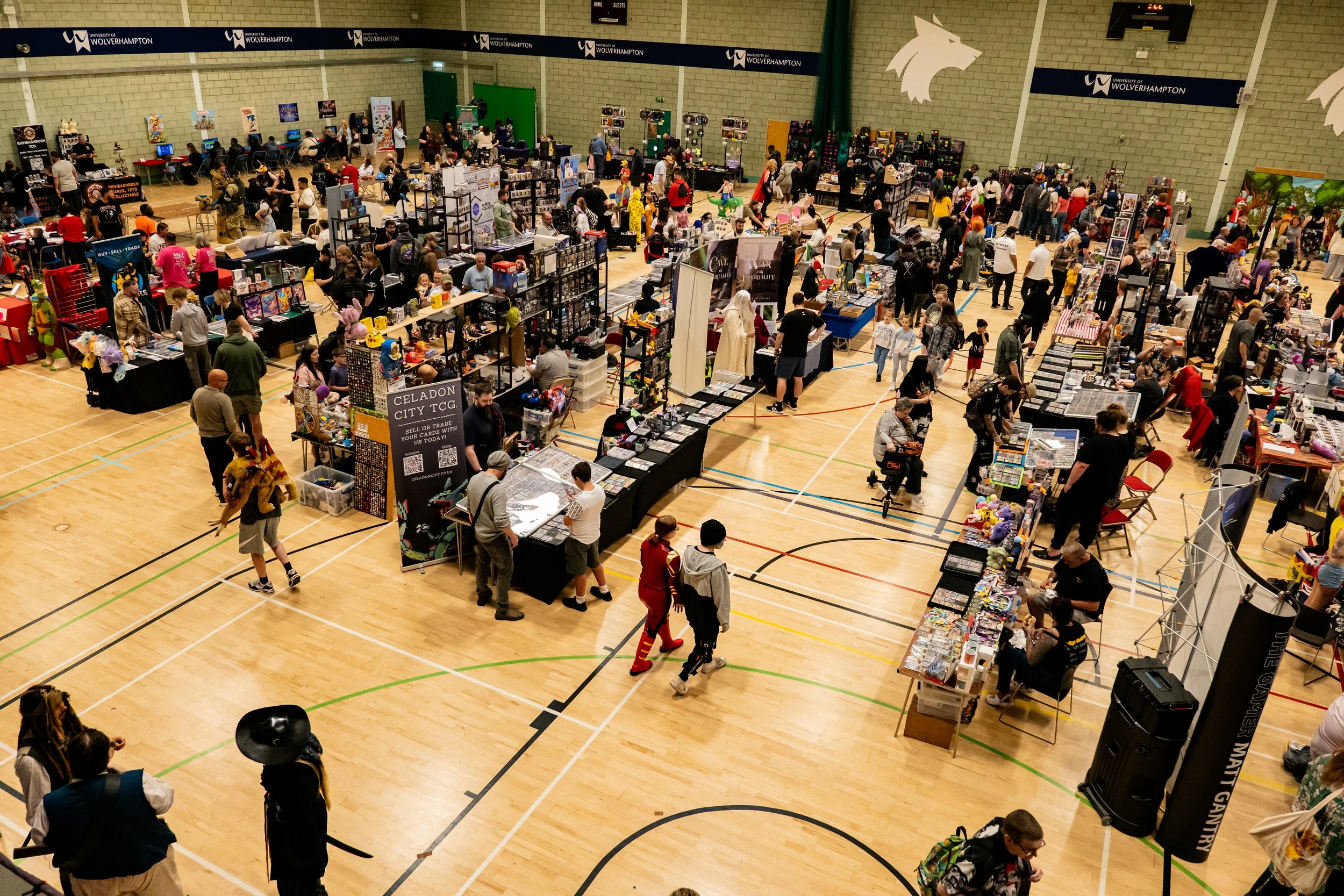 A large indoor convention with numerous tables and booths selling various collectibles, toys, and merchandise. Attendees are browsing and walking around, with some engaging with vendors. The venue is a gymnasium with a wooden floor, and there are ban