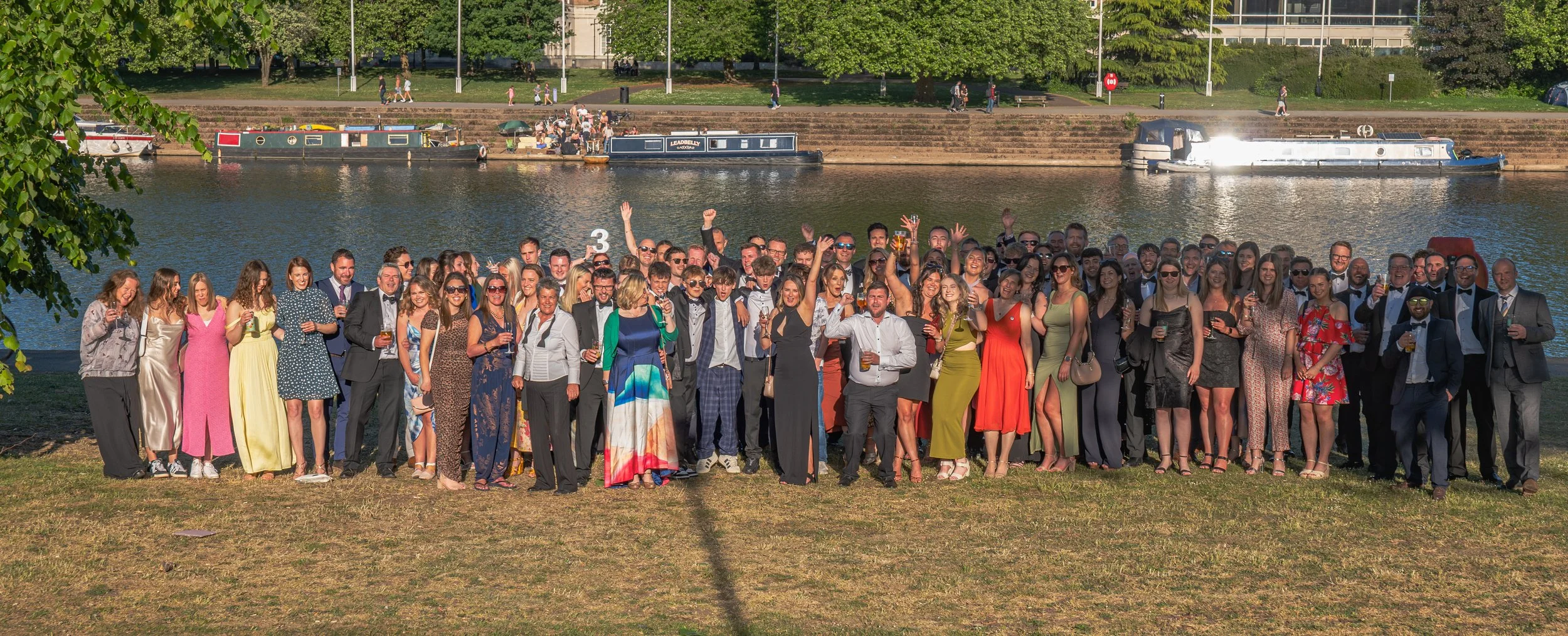 A large group of people dressed in formal attire, celebrating outdoors near a river with narrowboats in the background. The group is smiling, some holding drinks, and posing for a group photo in bright daylight.