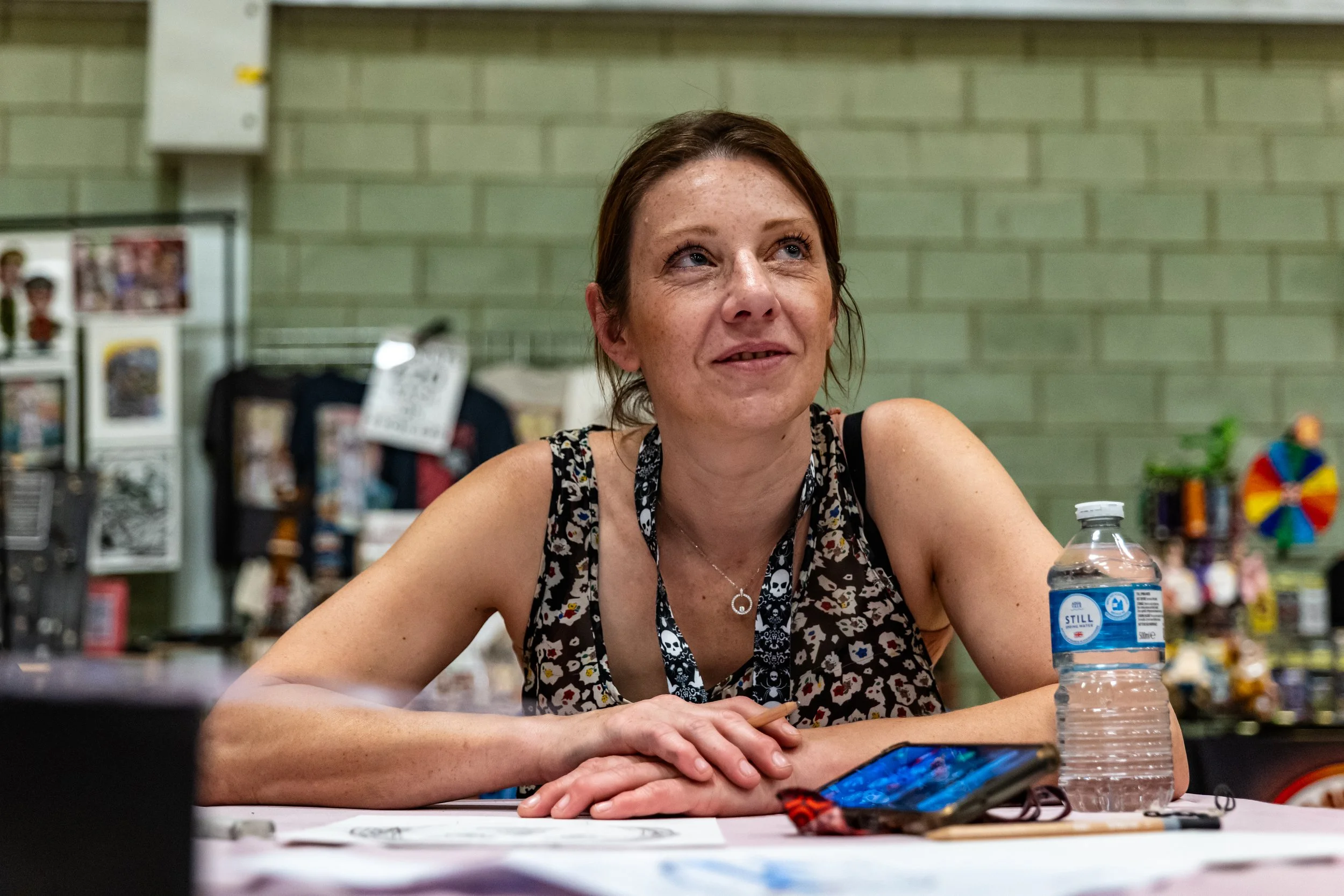 A woman with reddish brown hair, wearing a sleeveless patterned top and lanyard, sitting at a table with her hands folded, smiling slightly, in an indoor setting with a green brick wall and various items in the background.