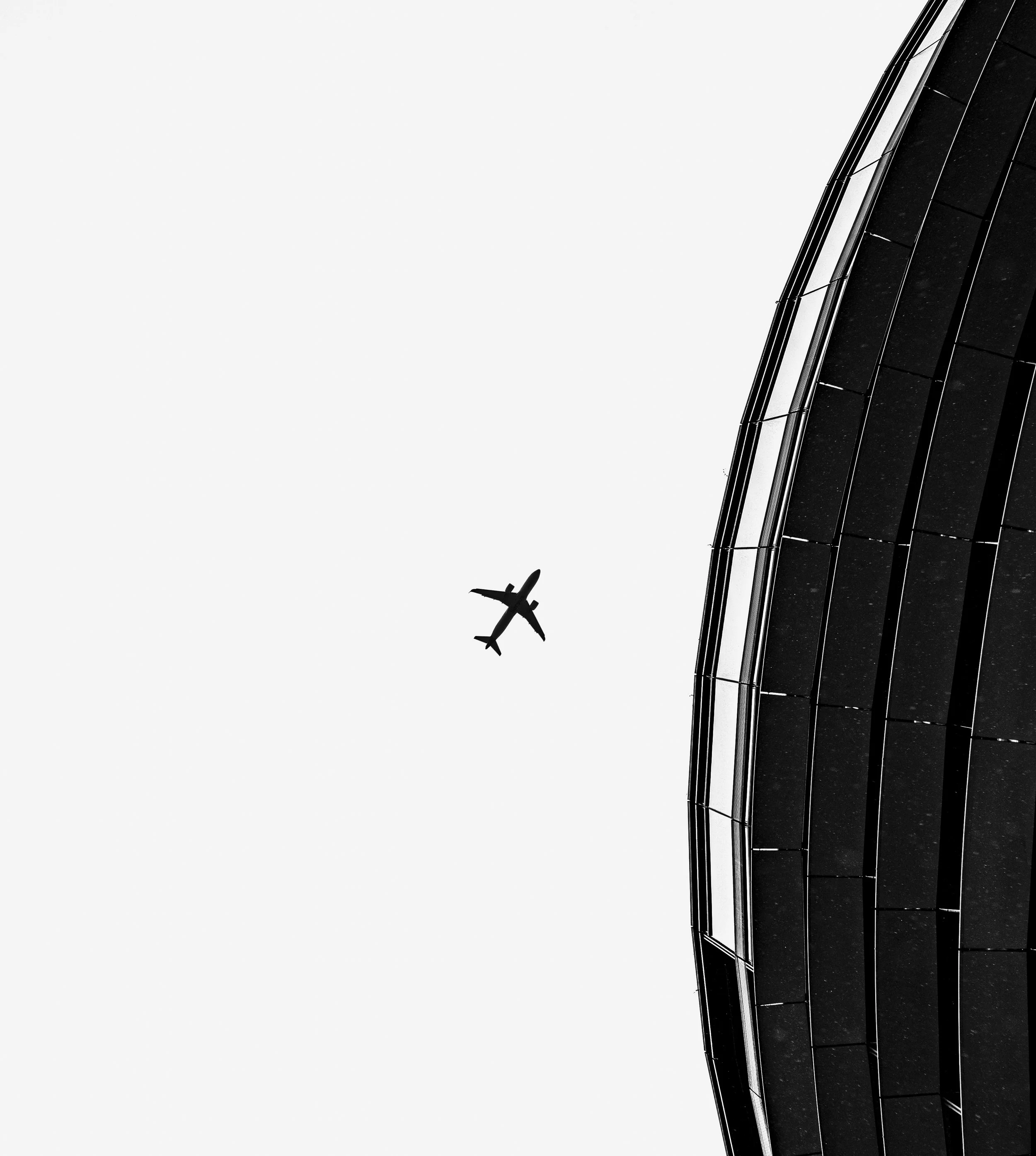 Silhouette of an airplane flying in the sky next to a modern glass building.