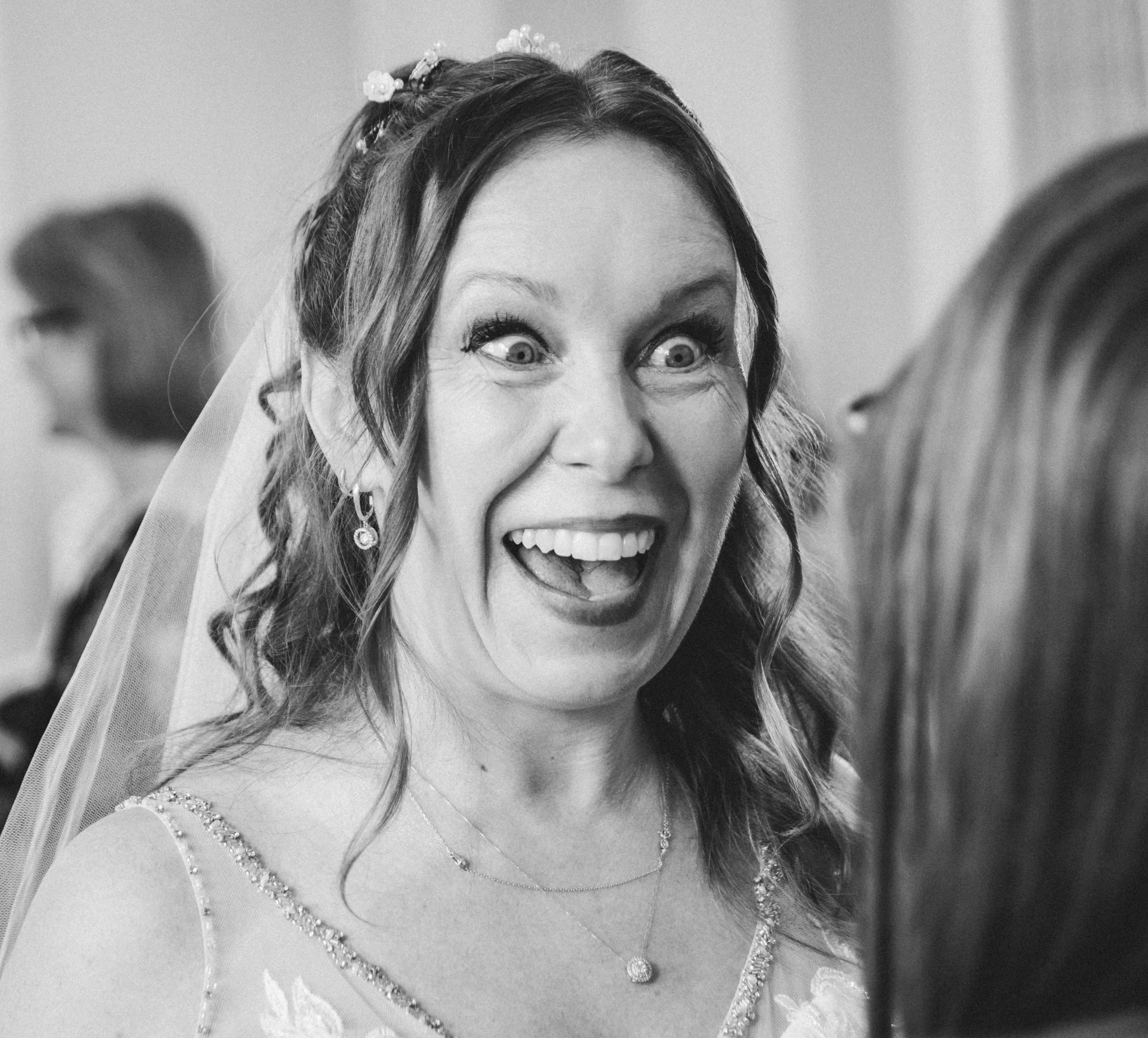 A woman with curly hair wearing a wedding dress, veil, and jewelry, making a surprised or excited facial expression.