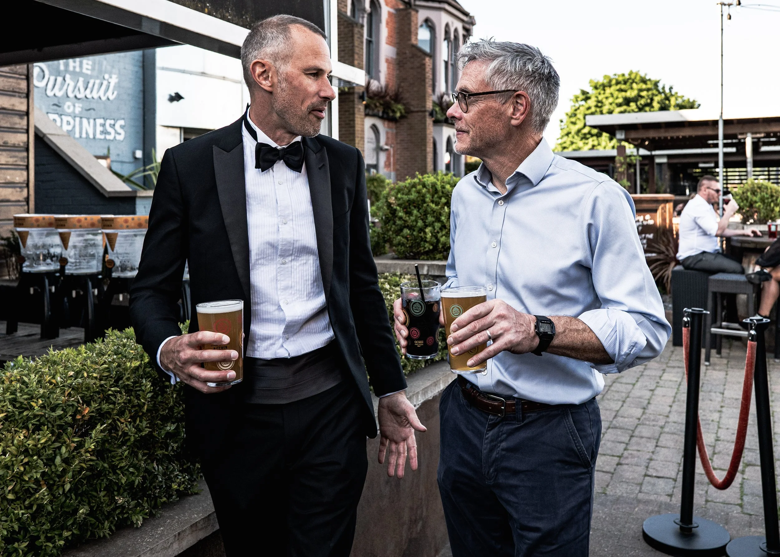 Two middle-aged men are conversing outdoors at a social gathering, holding drinks, one dressed in a tuxedo and the other in a button-down shirt and glasses, with a restaurant or bar in the background.