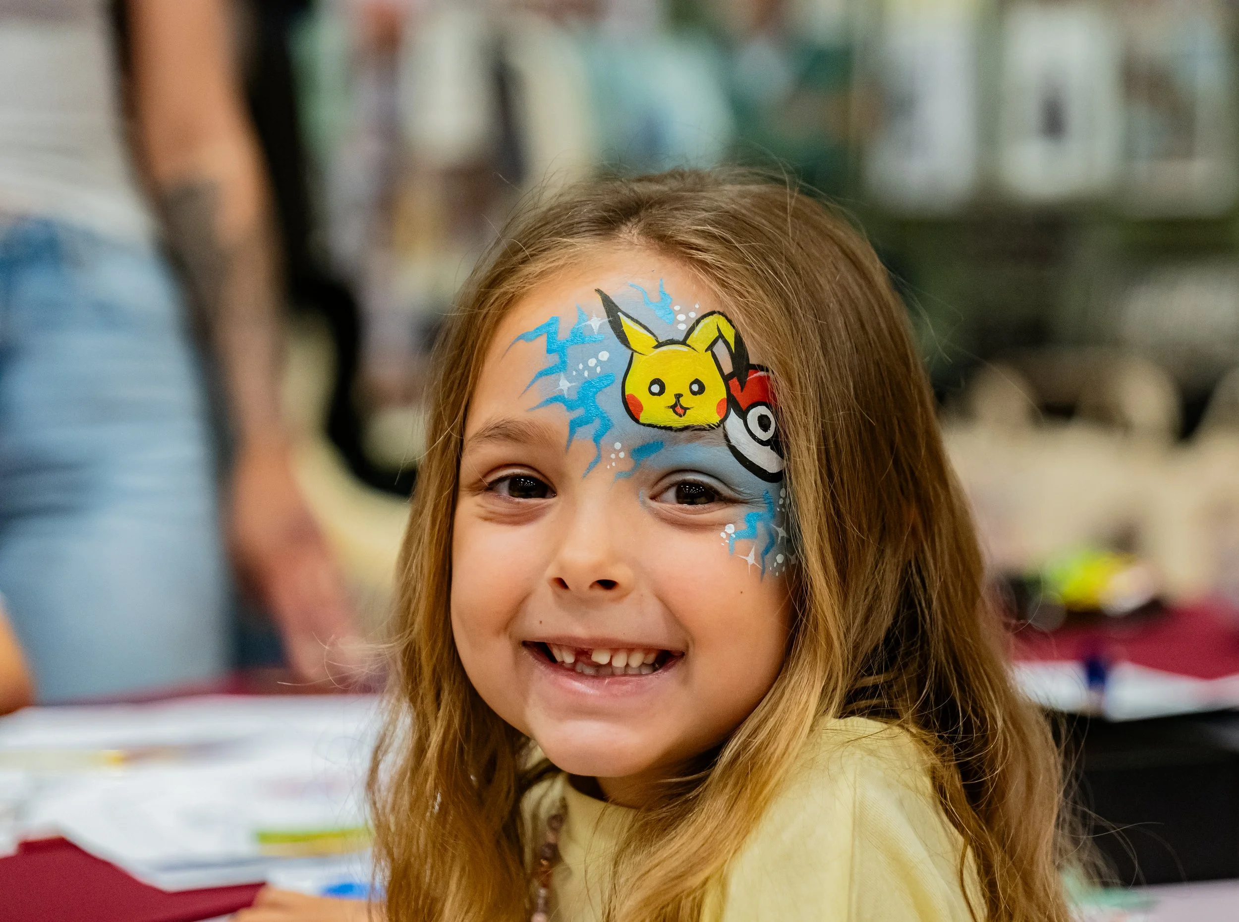 Young girl with face paint of Pikachu, Poké Ball, and lightning bolts, smiling at camera.
