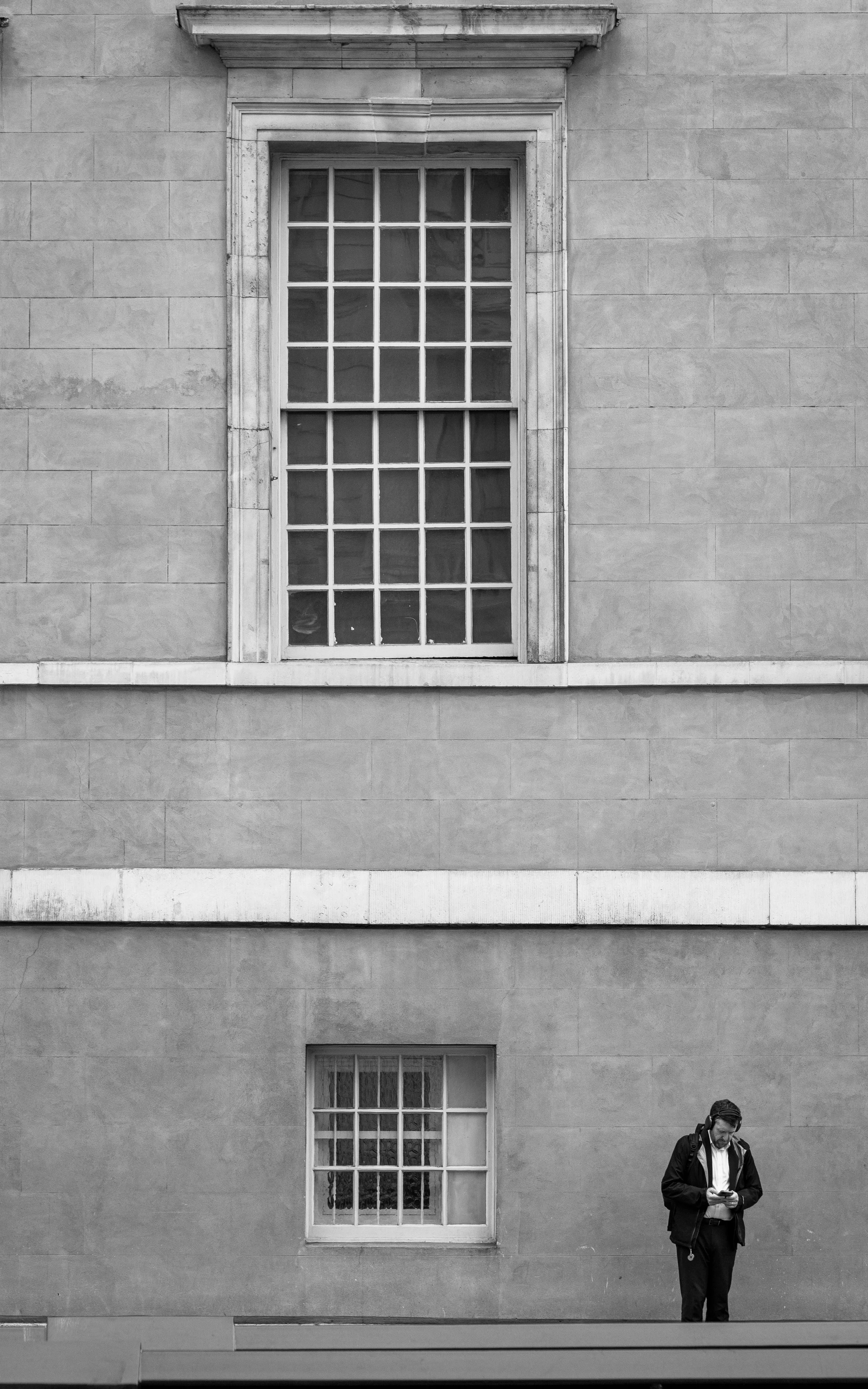 A man in a black jacket and slacks standing outside a building with a large window, looking at his phone.