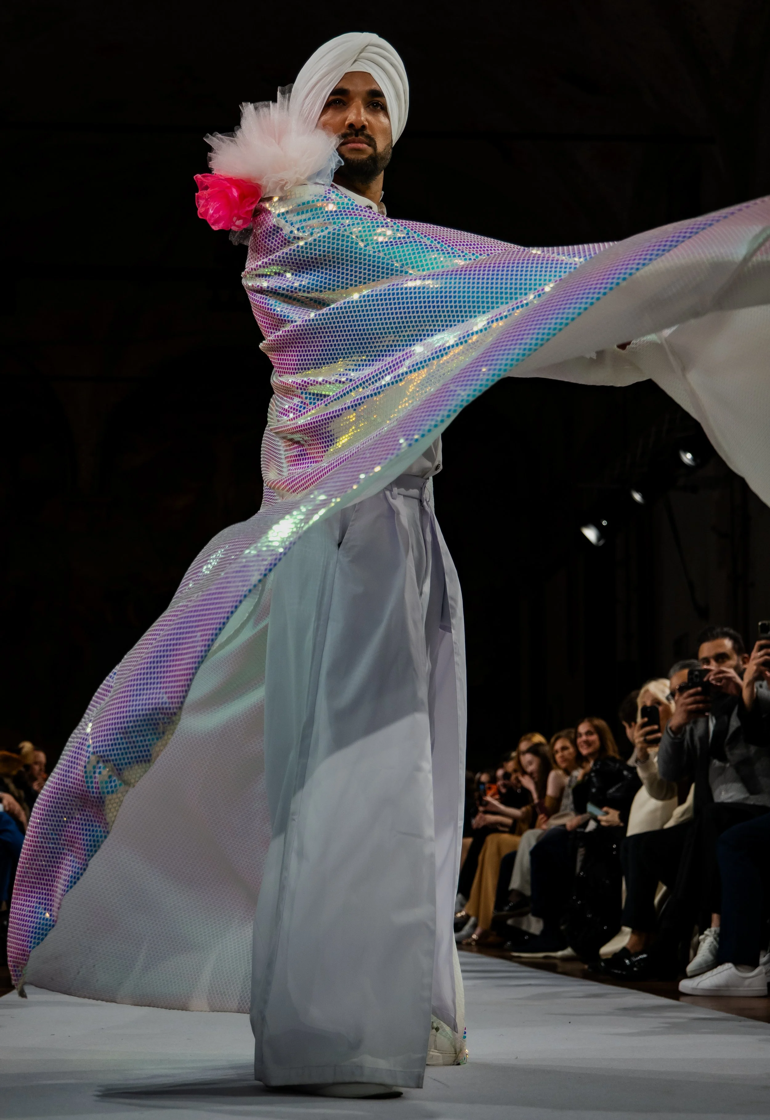 A male model wearing a white turban, flowing iridescent cape, and white traditional attire, walking on the runway at a fashion show with an audience taking photos.