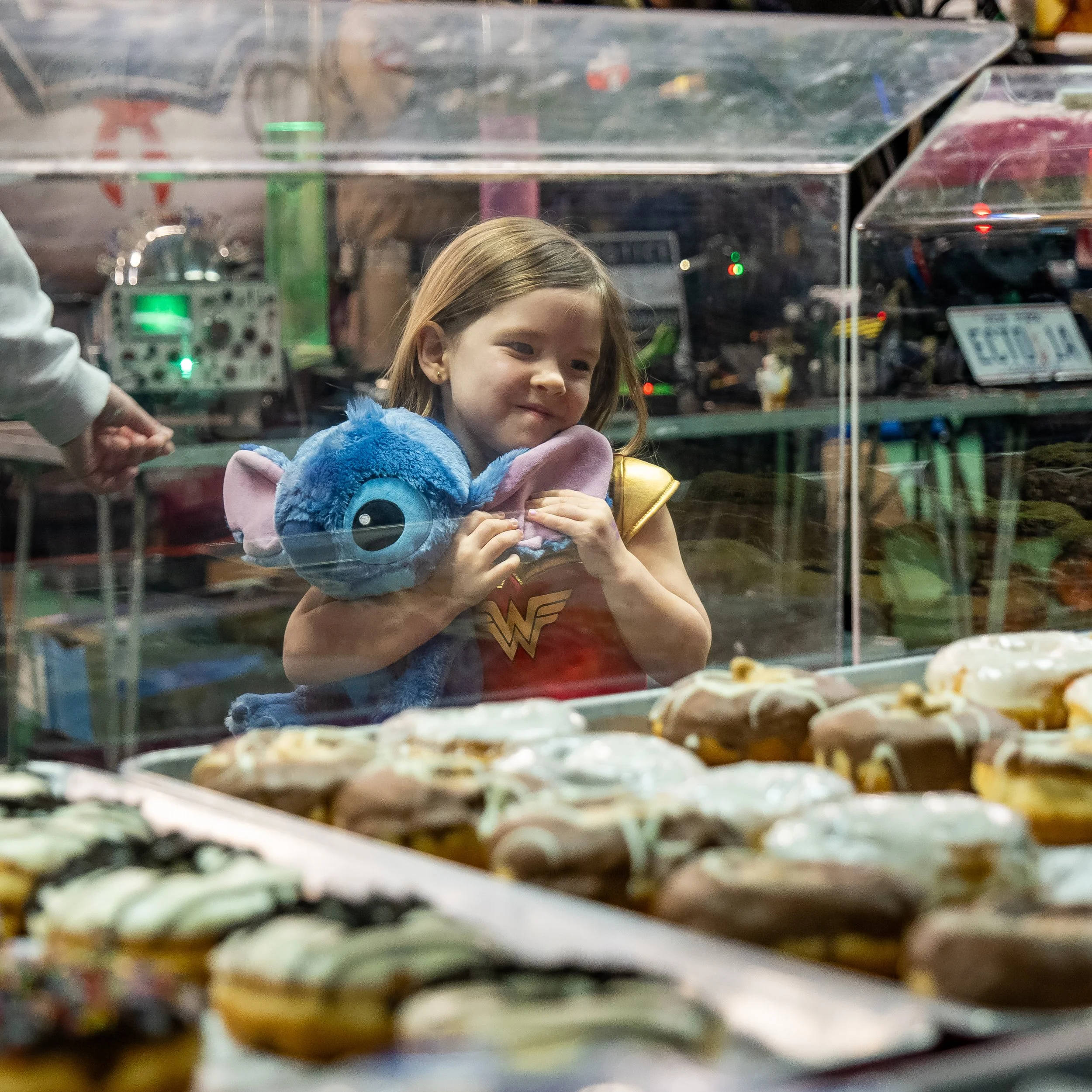 Young girl in Wonder Woman shirt holding a blue plush toy in front of a display case filled with donuts at a bakery or shop.
