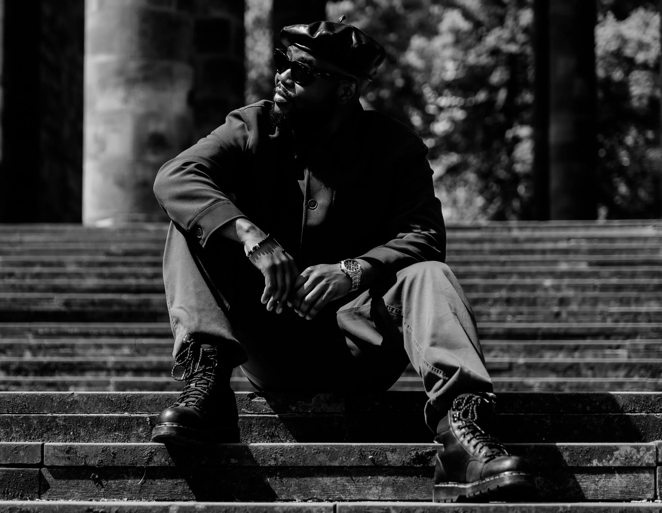 A man wearing sunglasses, a leather beret, a dark jacket, baggy pants, and combat boots, sitting on outdoor stairs with a blurred background of trees.