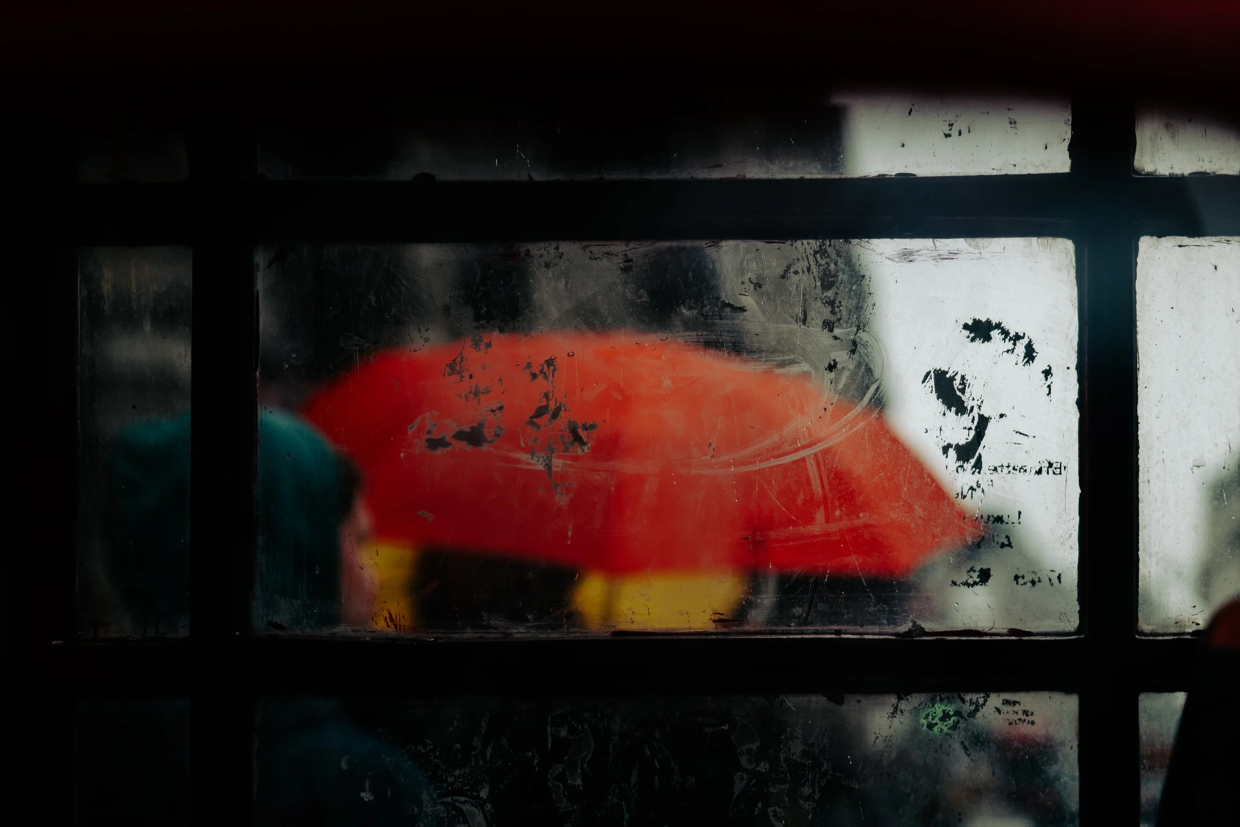Person holding a red umbrella, seen through a dirty window with black framing.