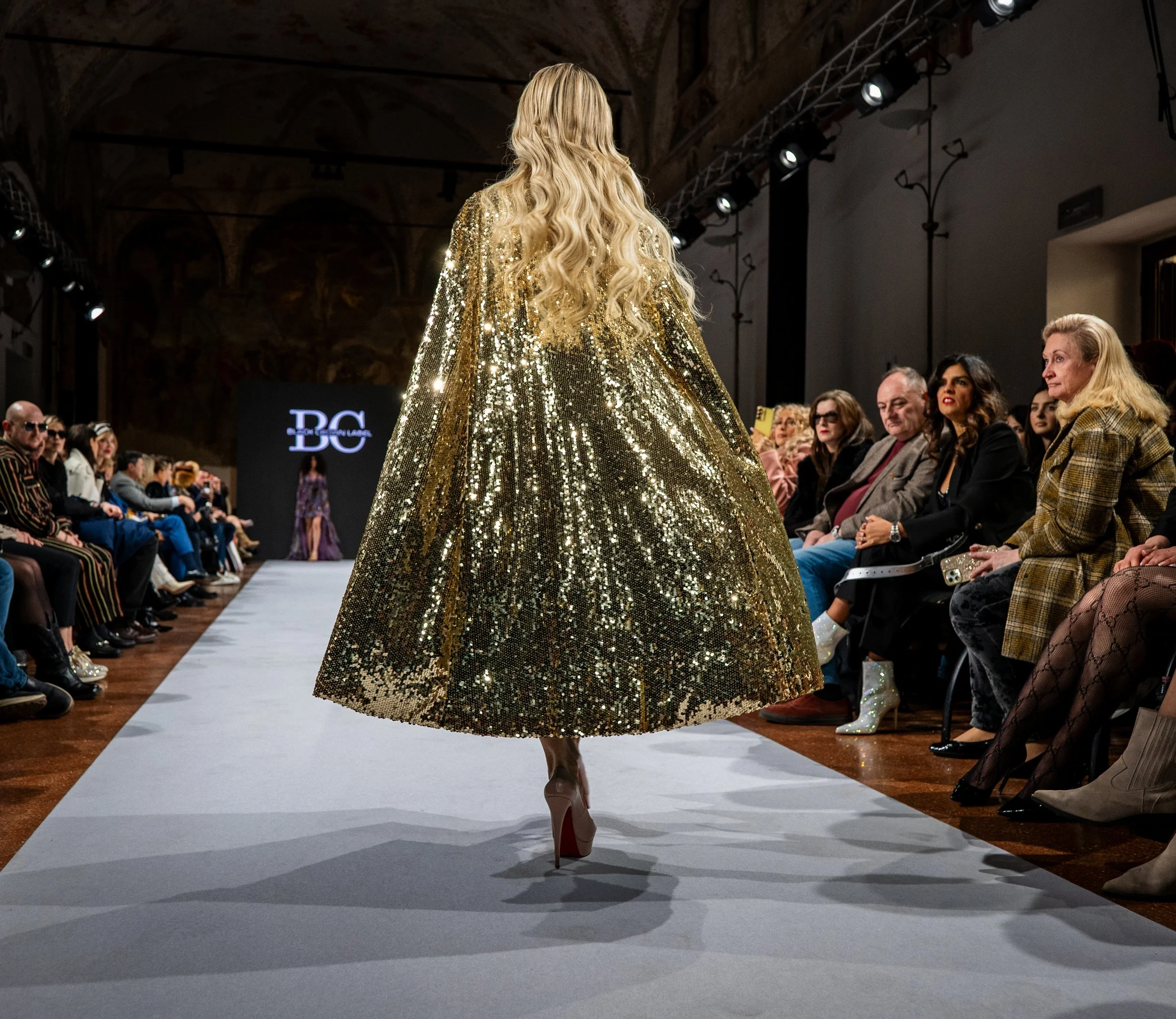 A model walking on a runway at a fashion show, wearing a large, sparkling gold sequin coat and high heels. Audience members are seated on both sides of the runway, watching the show.