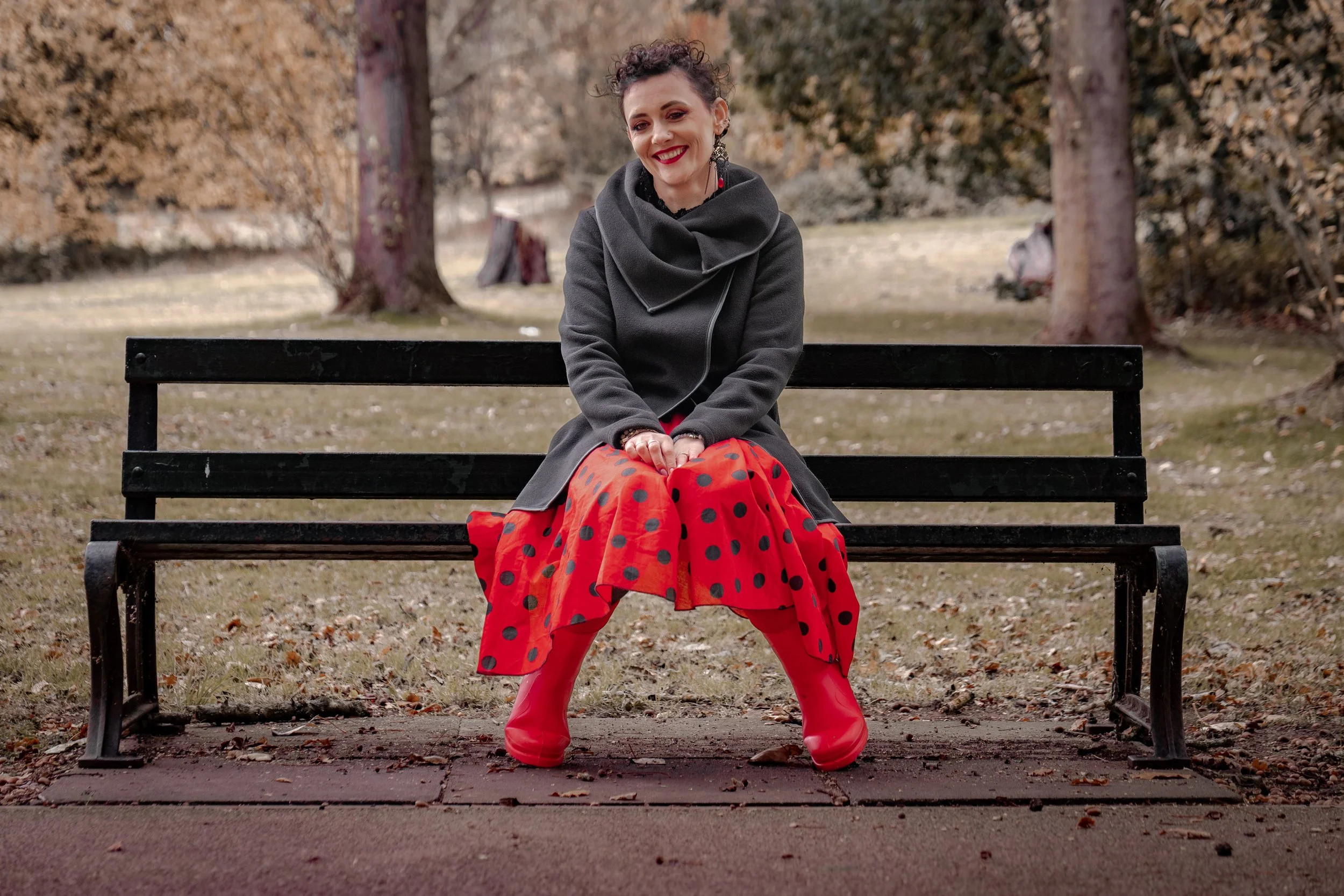 A woman sitting on a park bench with trees and grass in the background. She is wearing a dark gray coat, a red skirt with black polka dots, red rain boots, and has curly hair and red lipstick, smiling at the camera.