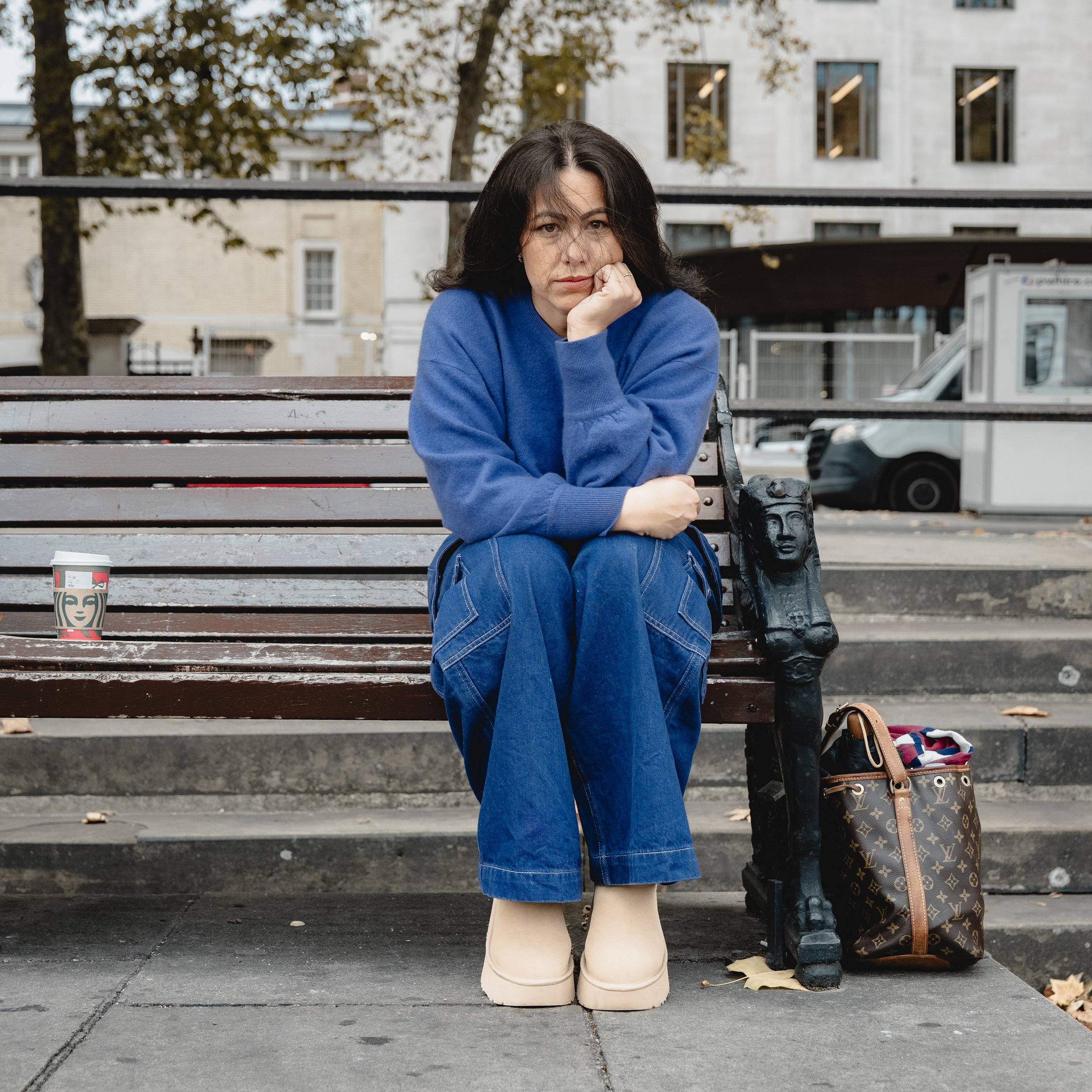 A woman sitting on a park bench with her head resting on her hand, wearing a blue sweater, blue jeans, and beige boots. There is a Starbucks cup on the bench next to her and a Louis Vuitton bag on the ground beside her. In the background, there are b