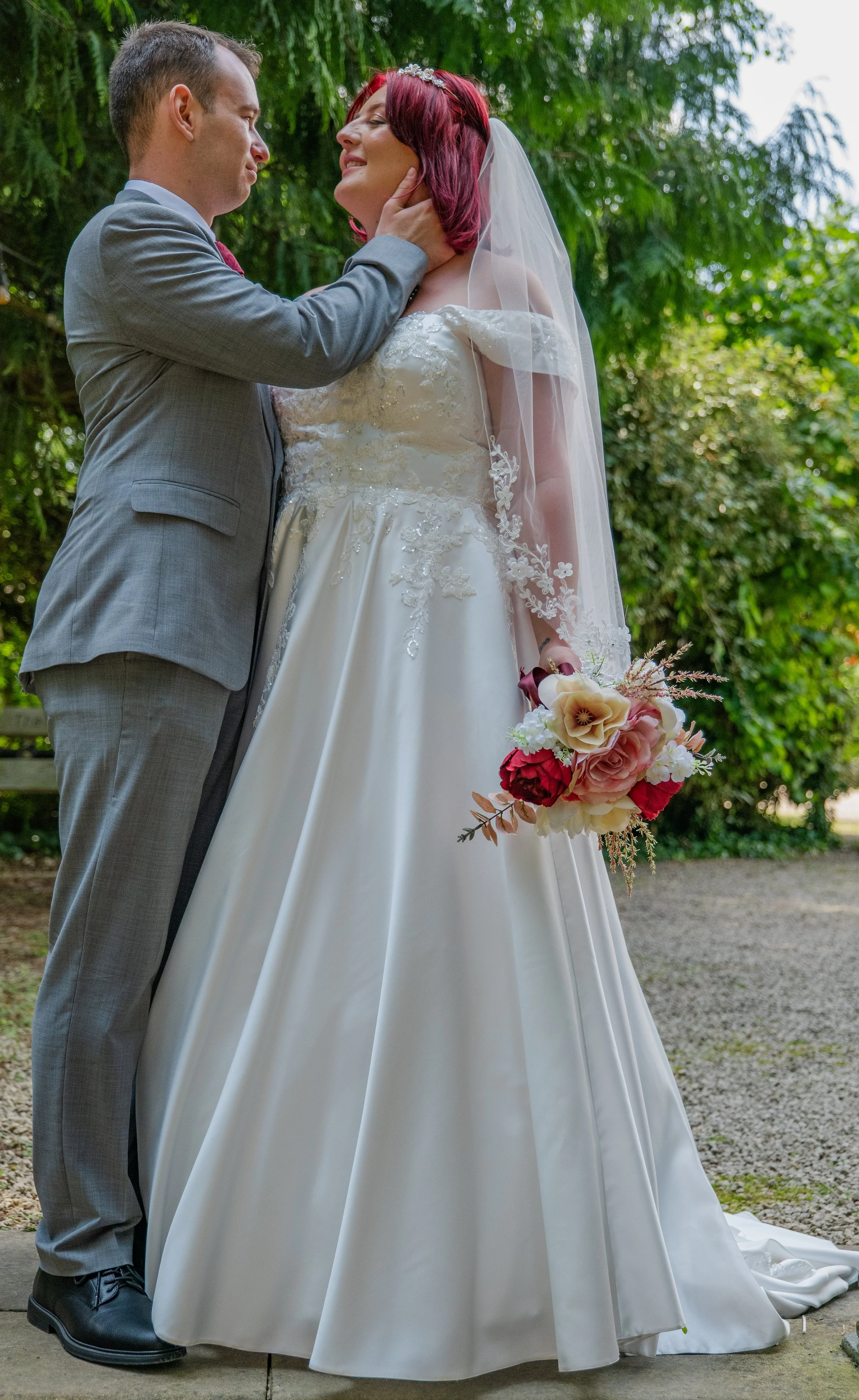 A bride and groom share an intimate moment outdoors, with trees in the background. The bride wears a white wedding gown with lace details, a veil, and holds a bouquet of flowers. The groom is dressed in a gray suit and is gently touching the bride's 