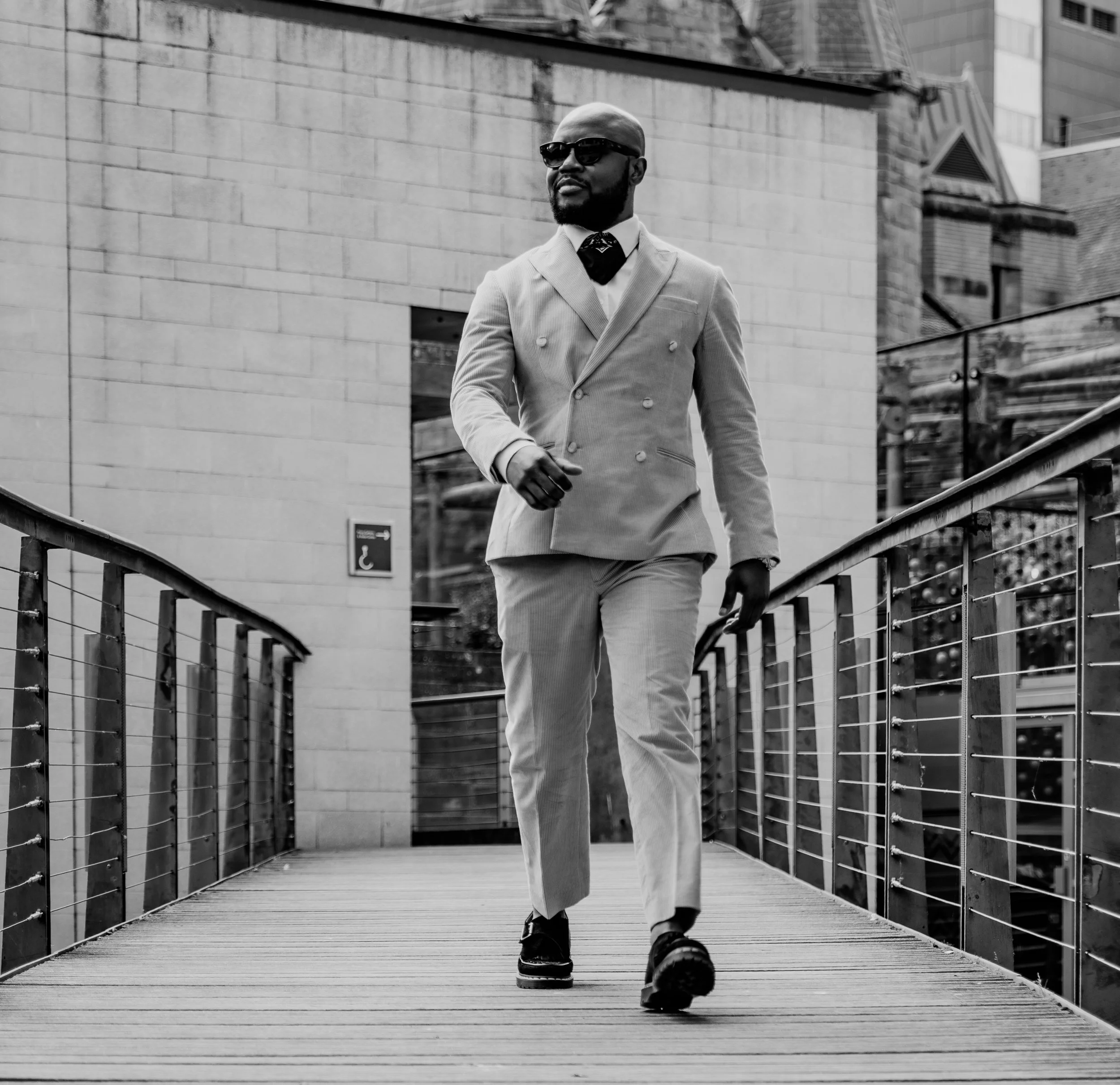 A stylish man in a suit wearing sunglasses walks across a bridge in an urban area, captured in black and white.