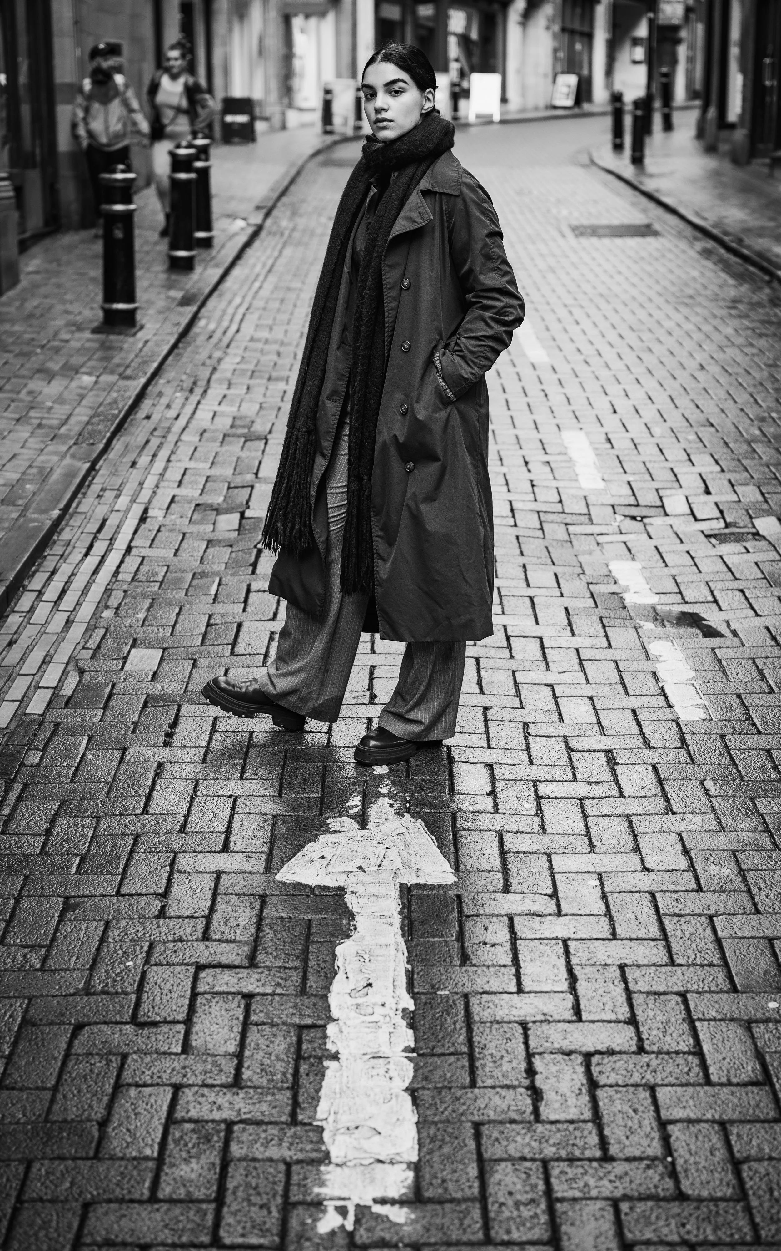 A woman with dark hair wearing a long coat, scarf, and dress, stands on a brick street with her hands in her coat pockets, looking at the camera.