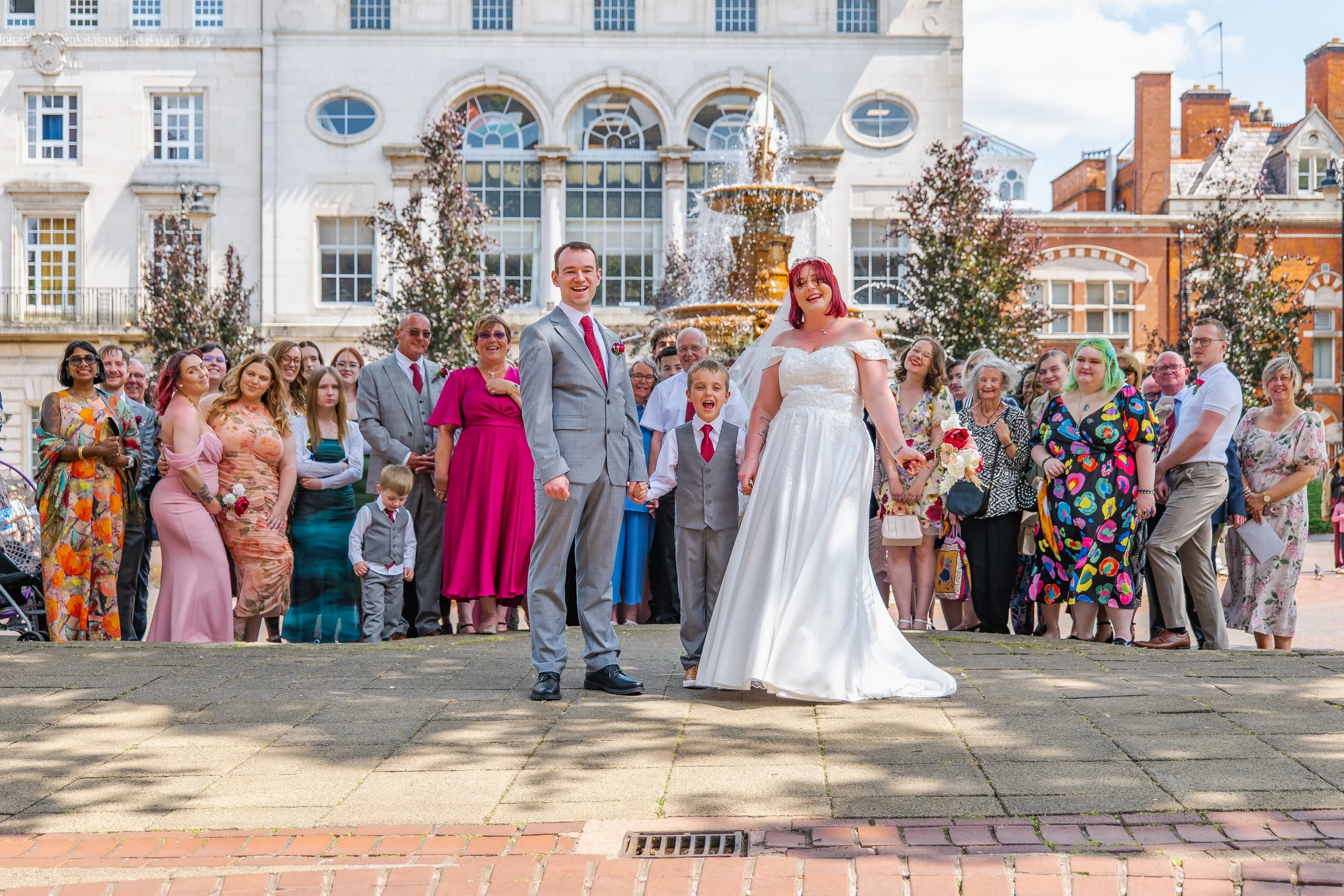 A wedding celebration with a bride and groom holding hands, surrounded by friends and family outdoors in front of a fountain and historic building.