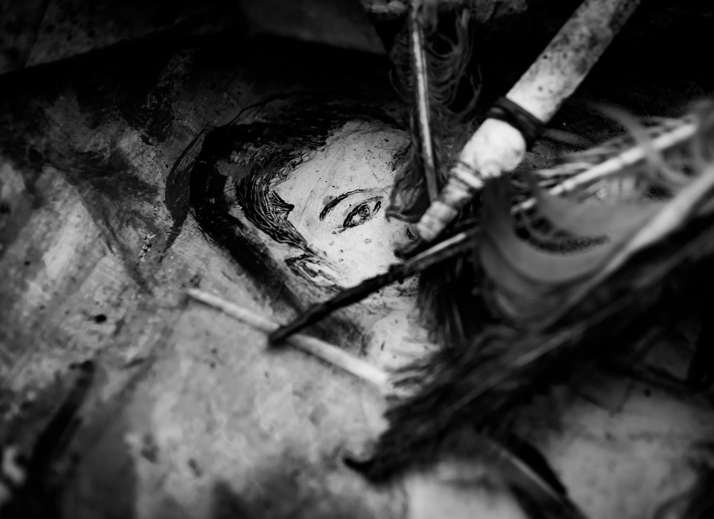 A black-and-white photo of a mural or painting of a woman's face being created on a surface, with brushes and painting tools around.