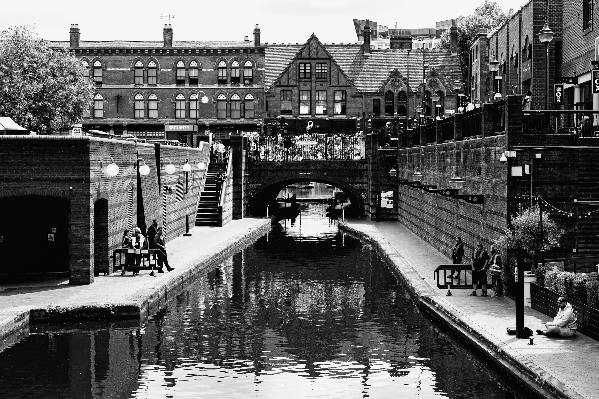 Black and white photo of a city canal with brick walls and a bridge, pedestrians walking on the sidewalk, buildings and businesses in the background.
