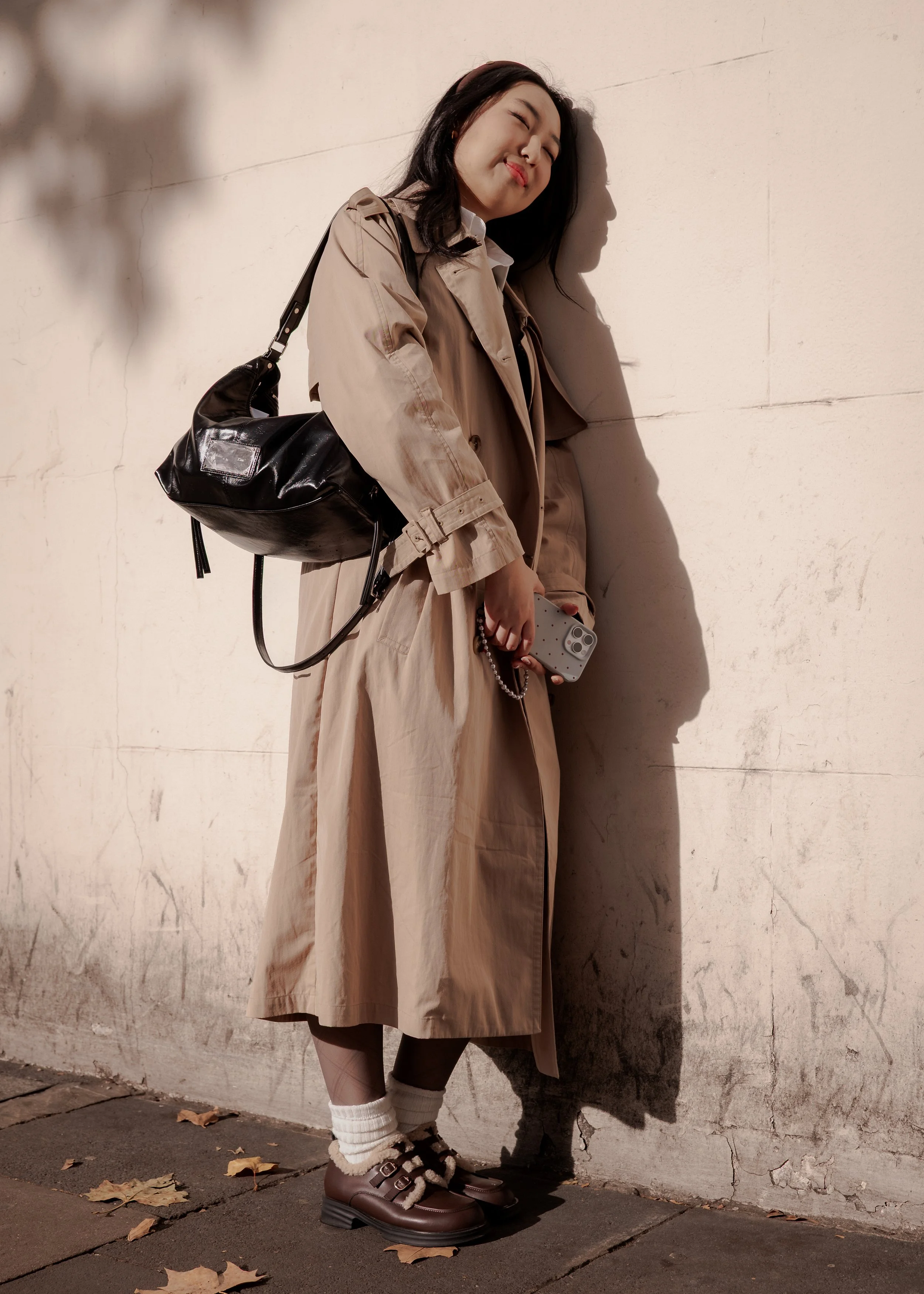 A young woman leans against a beige wall, smiling with eyes closed, holding a smartphone. She is dressed in a beige trench coat, brown shoes, and white socks, carrying a black bag over her shoulder. Shadows and fallen leaves are visible on the paveme