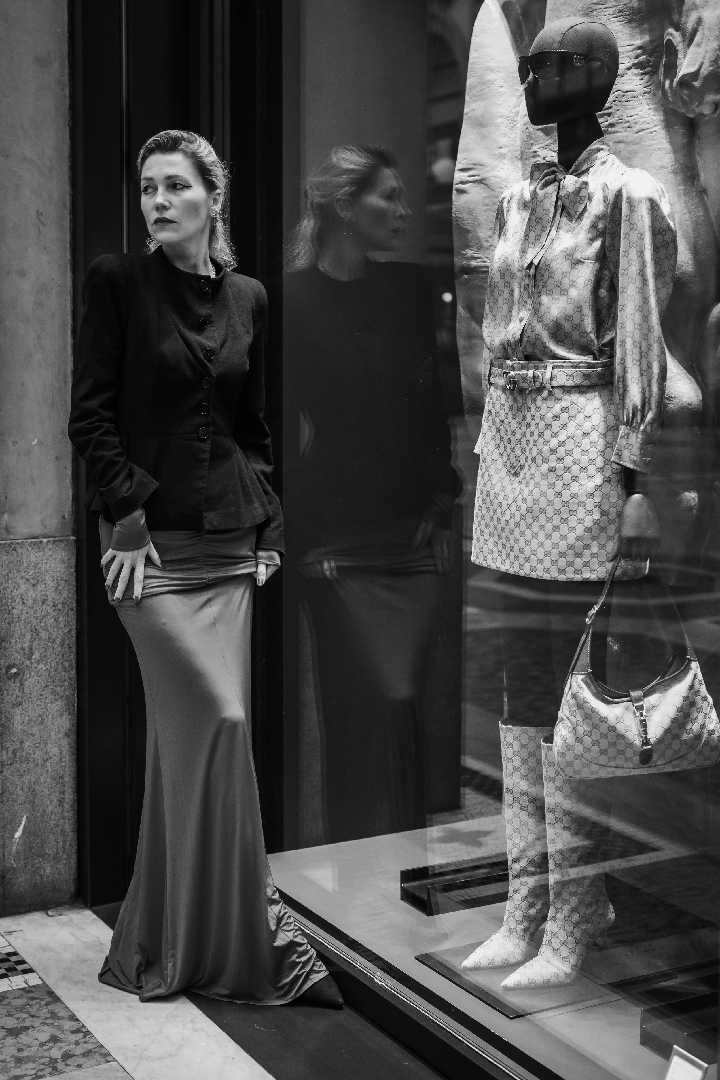 Woman standing outside a clothing store window, looking to her right. The store display includes a mannequin dressed in designer clothing and accessories, with her reflection visible in the glass.
