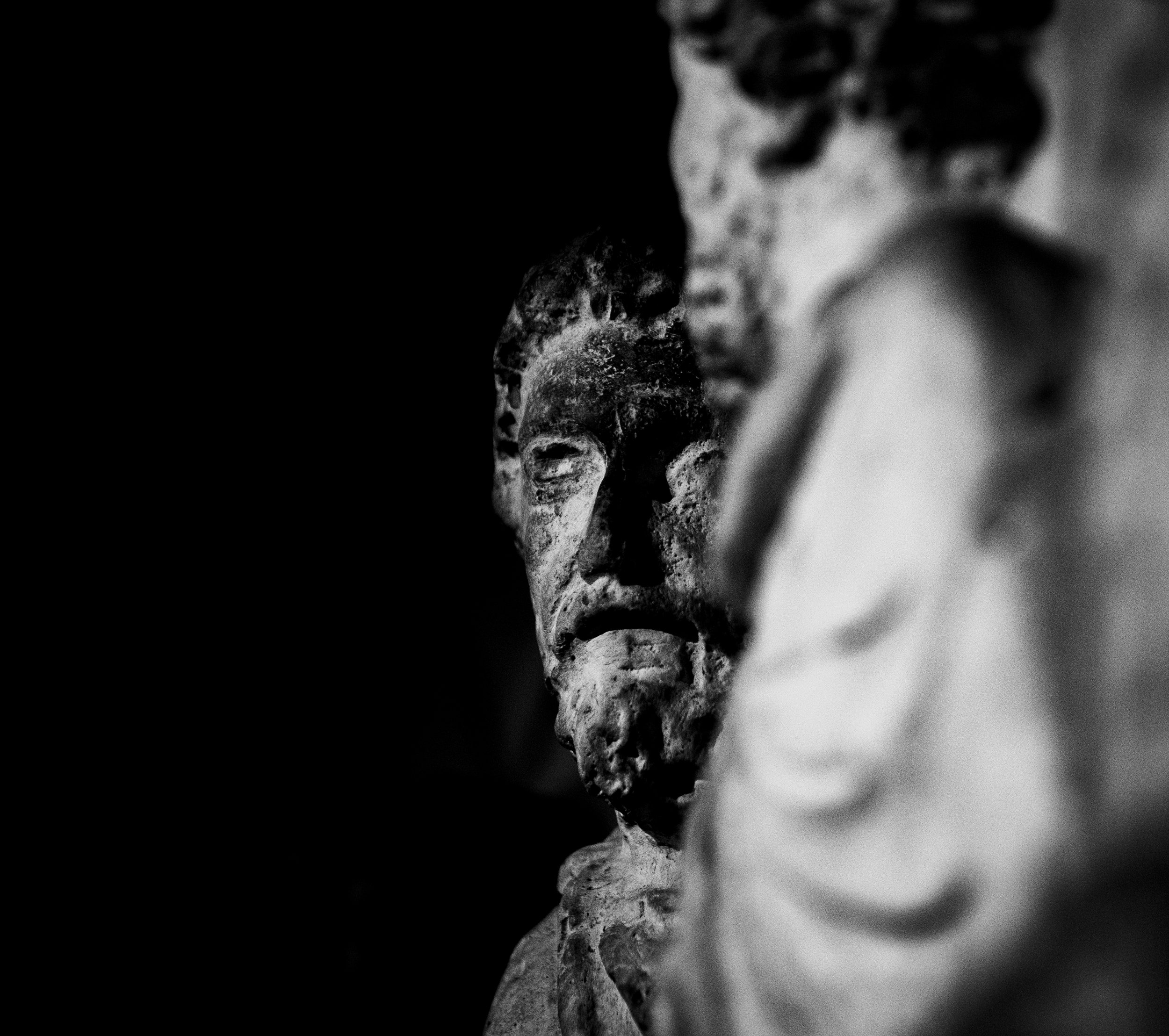 Close-up of weathered stone sculptures, with one face visible, set against a dark background, emphasizing texture and shadows in black and white.