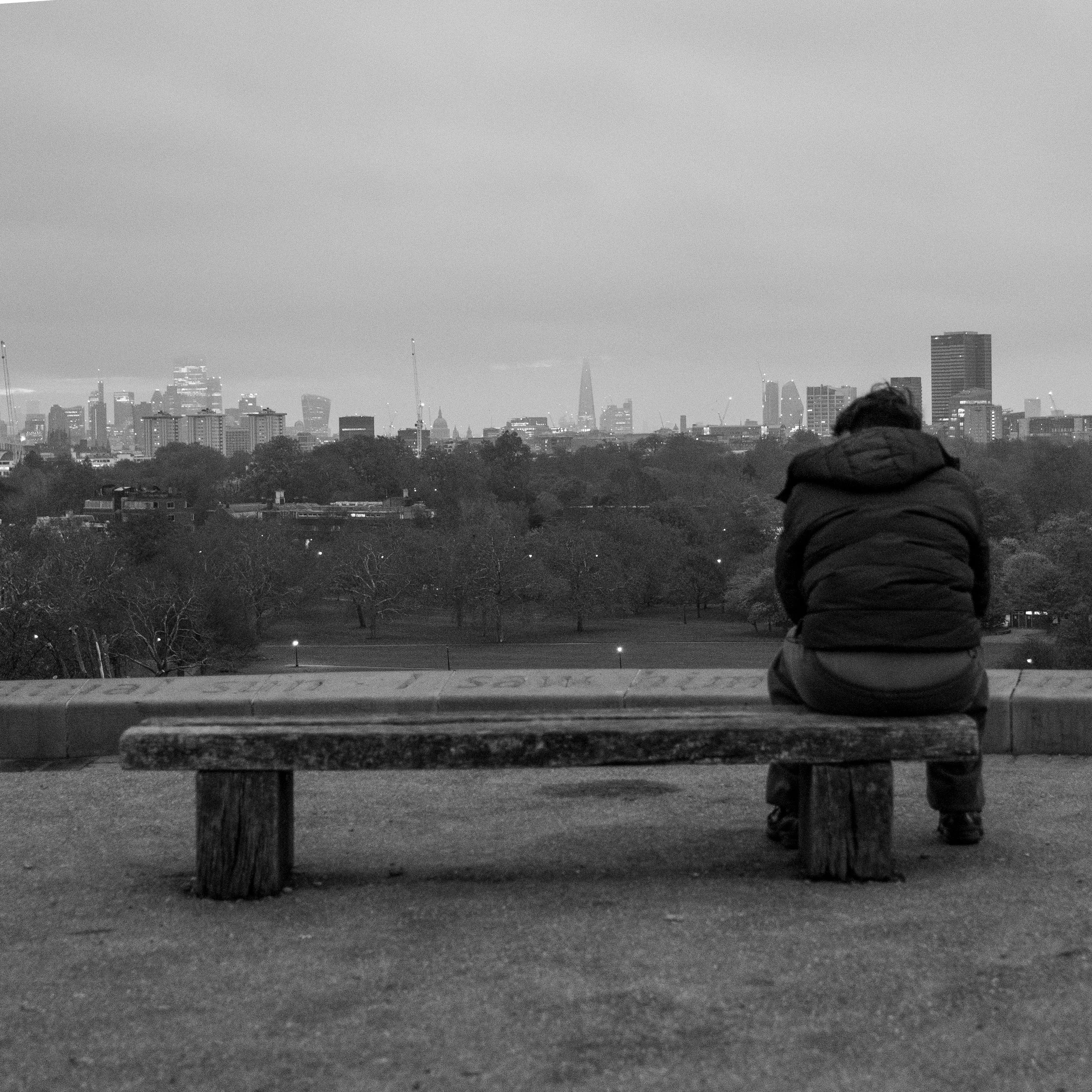 Person sitting alone on a wooden bench in a park, looking over a city skyline in the distance, in black and white.