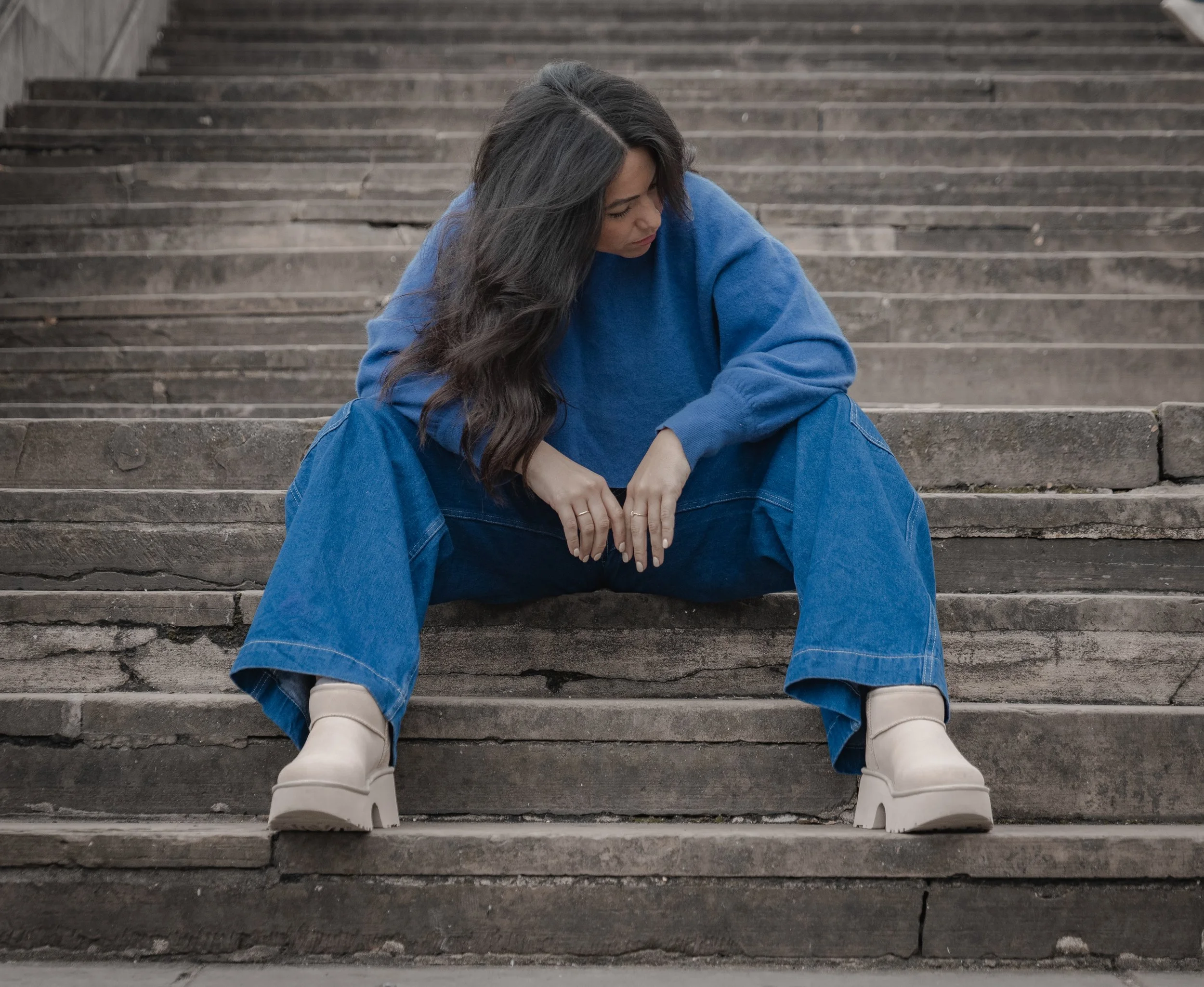 Woman in a blue sweatshirt and jeans sitting on outdoor stone steps, looking down with her arms resting on her knees.