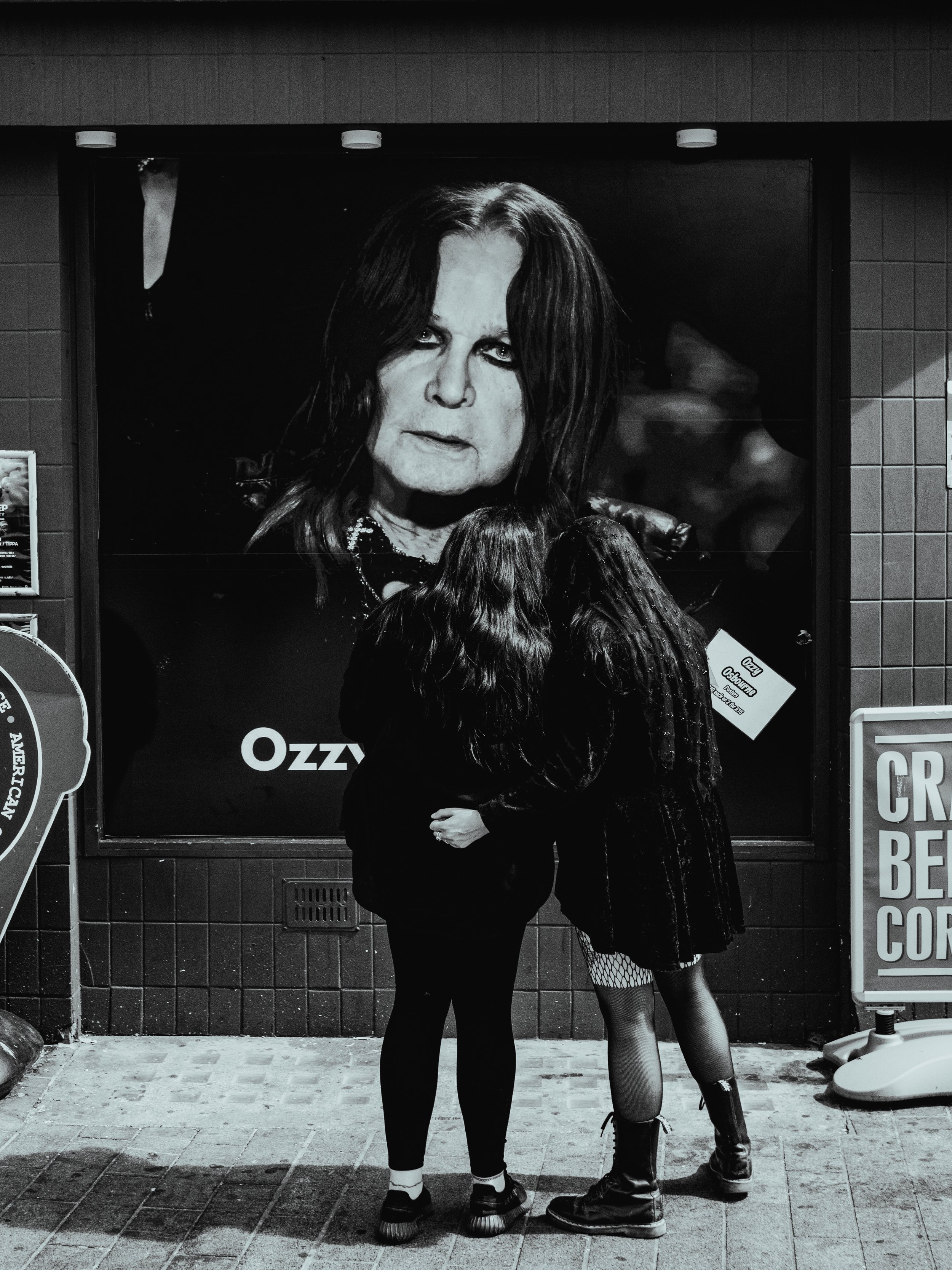 Two women with dark hair, dressed in dark clothing, standing close together and kissing outside a building at night. A large poster of Ozzy Osbourne is behind them.