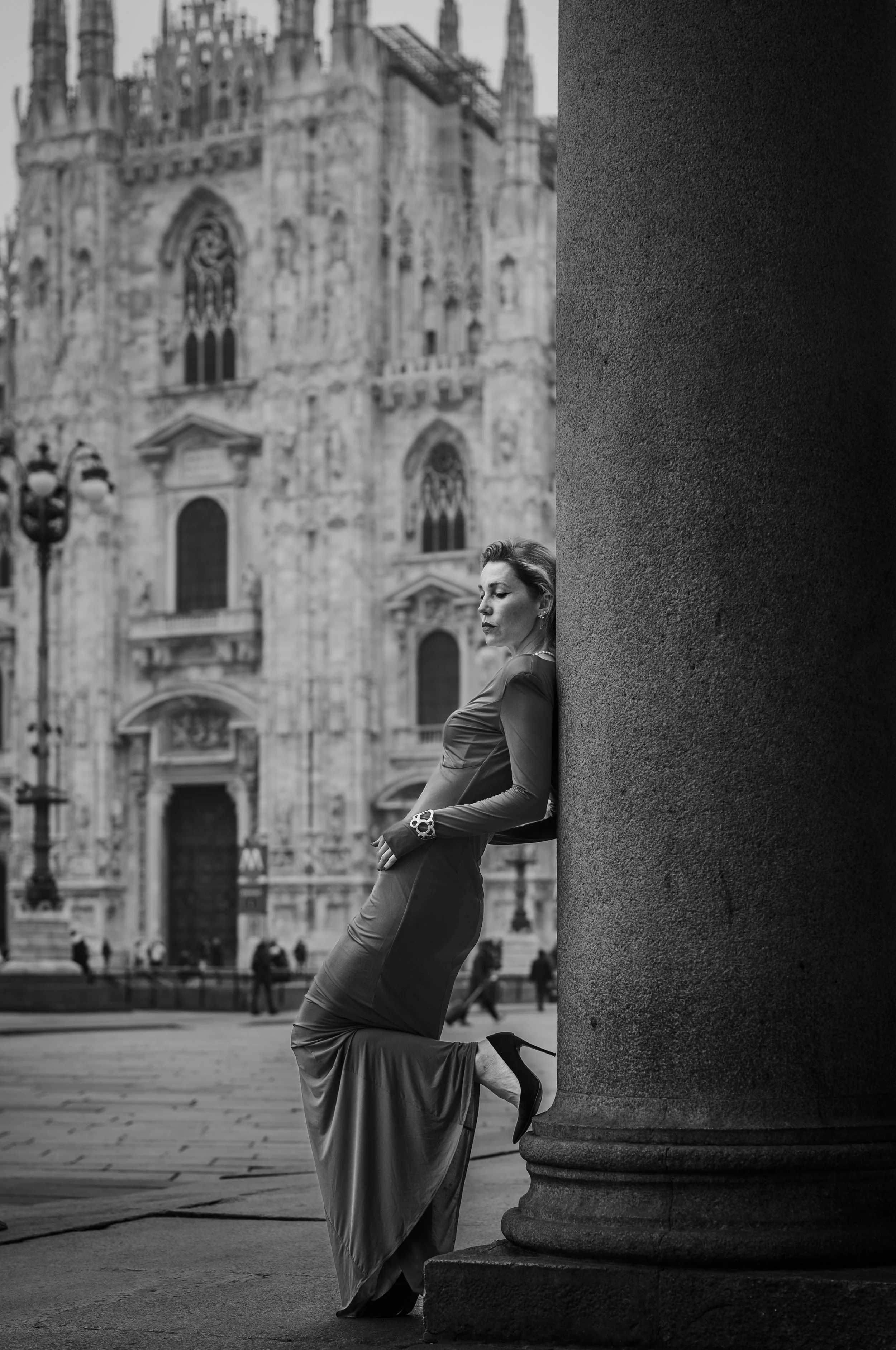 A woman in a long dress and high heels leaning against a large column in front of an ornate historic building.