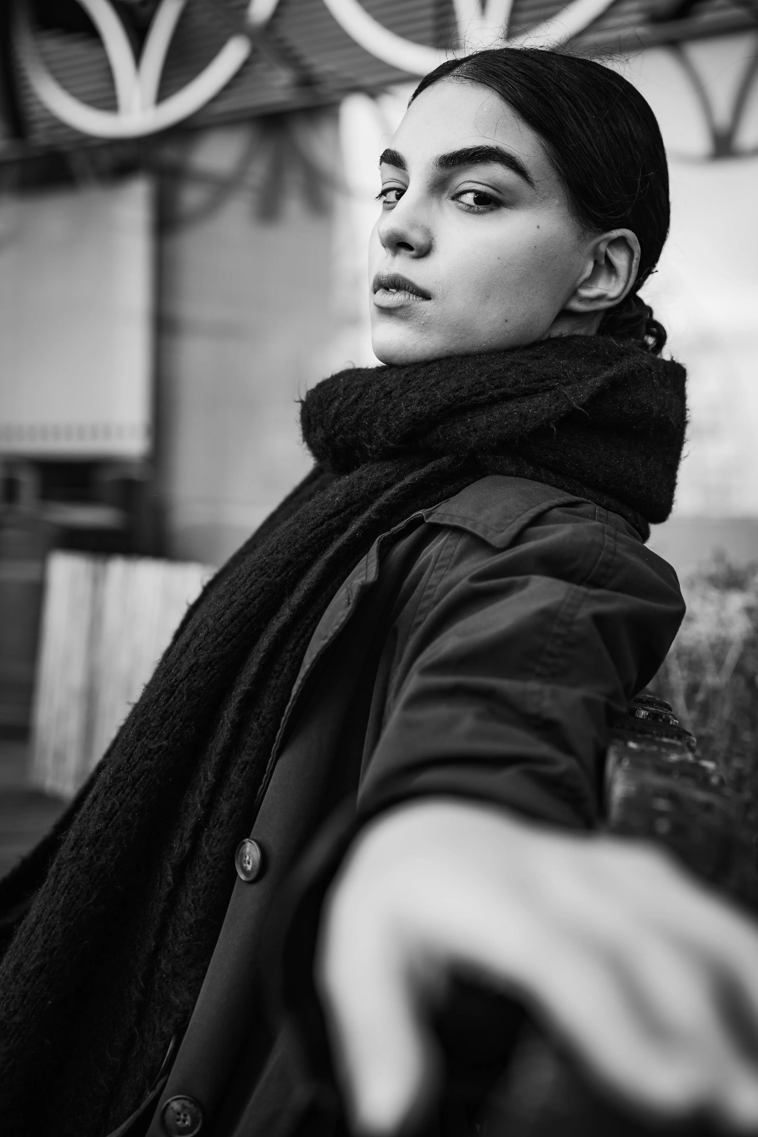 Black and white portrait of a young woman with dark hair pulled back, wearing a scarf and jacket, looking over her shoulder with a confident expression.