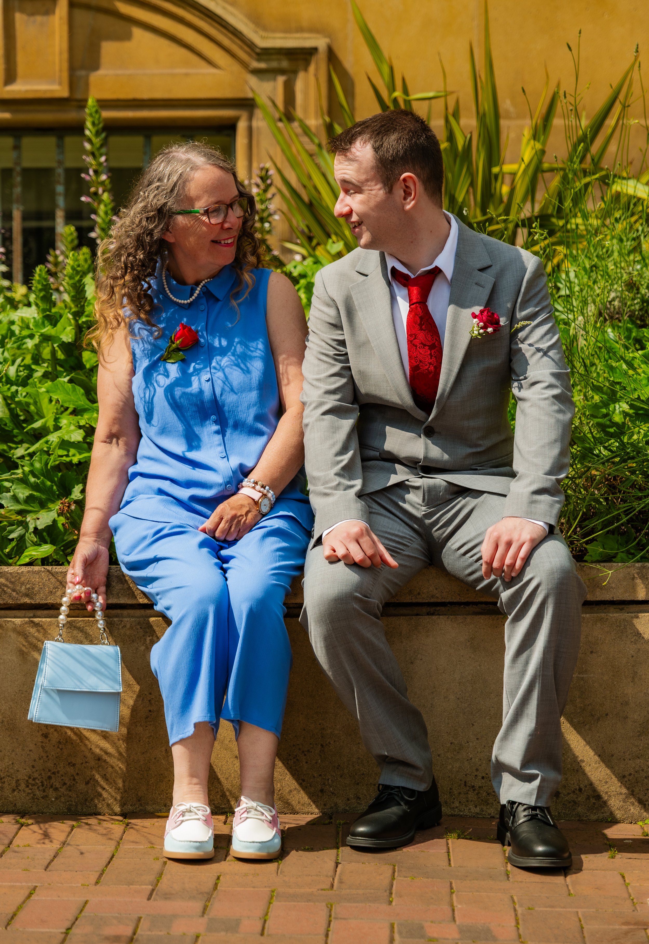 A woman in a blue dress and a man in a gray suit sit and smile at each other in a garden setting.