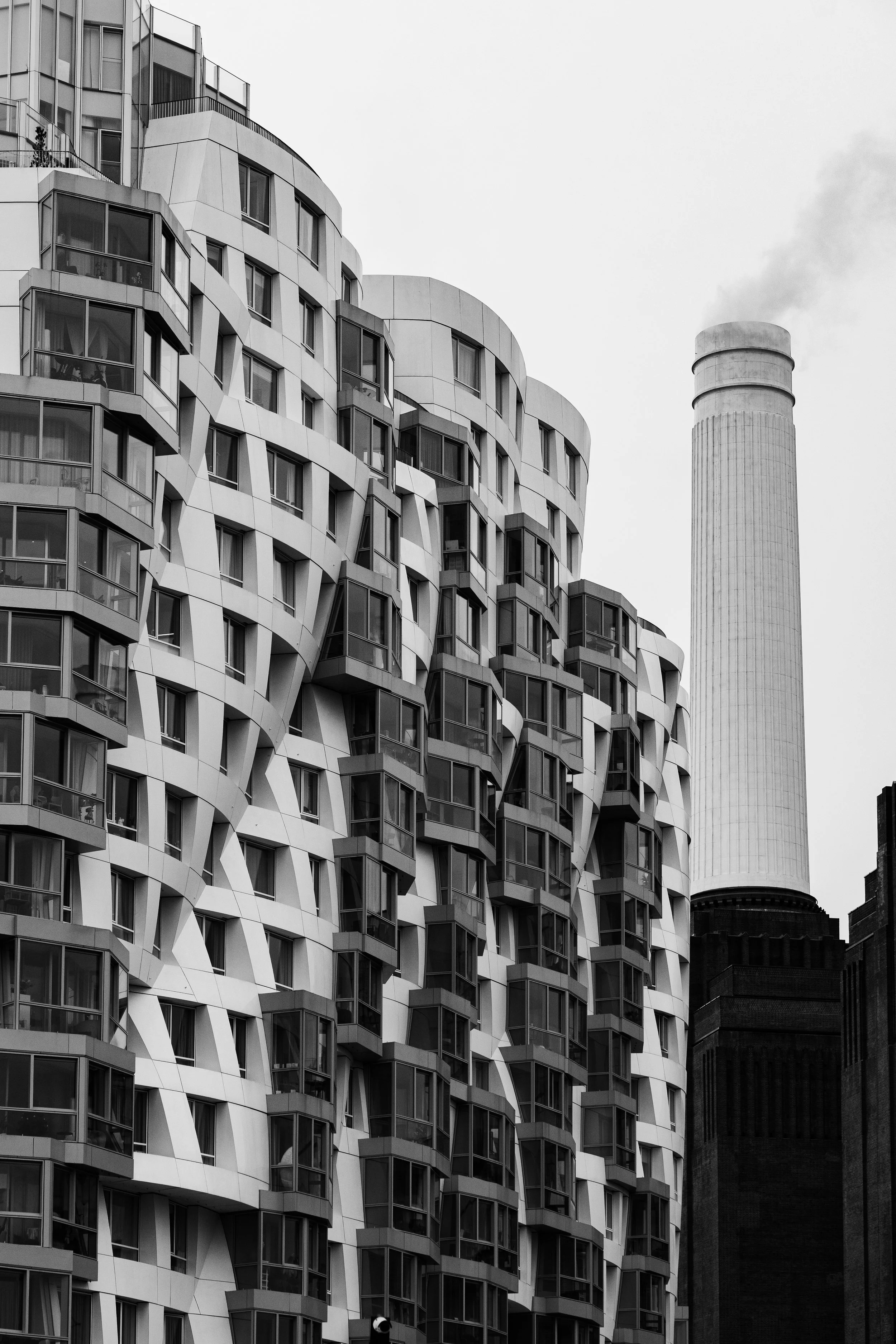 A black and white photo of a modern apartment building with a wavy facade and multiple balconies, with a tall industrial chimney emitting smoke in the background.