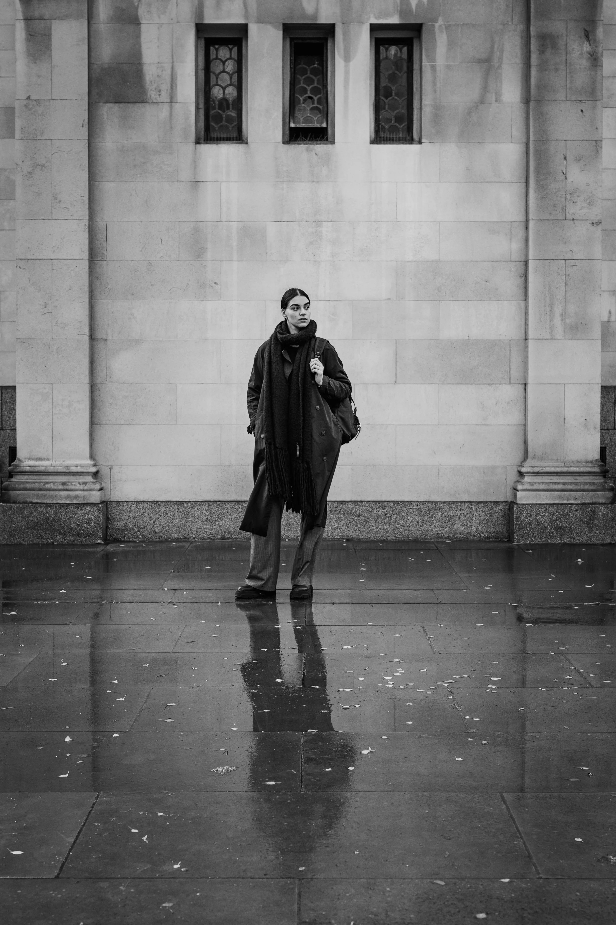 A woman in a dark coat and scarf standing on a wet city sidewalk, reflected in puddles, in front of a stone building with four small, patterned windows.